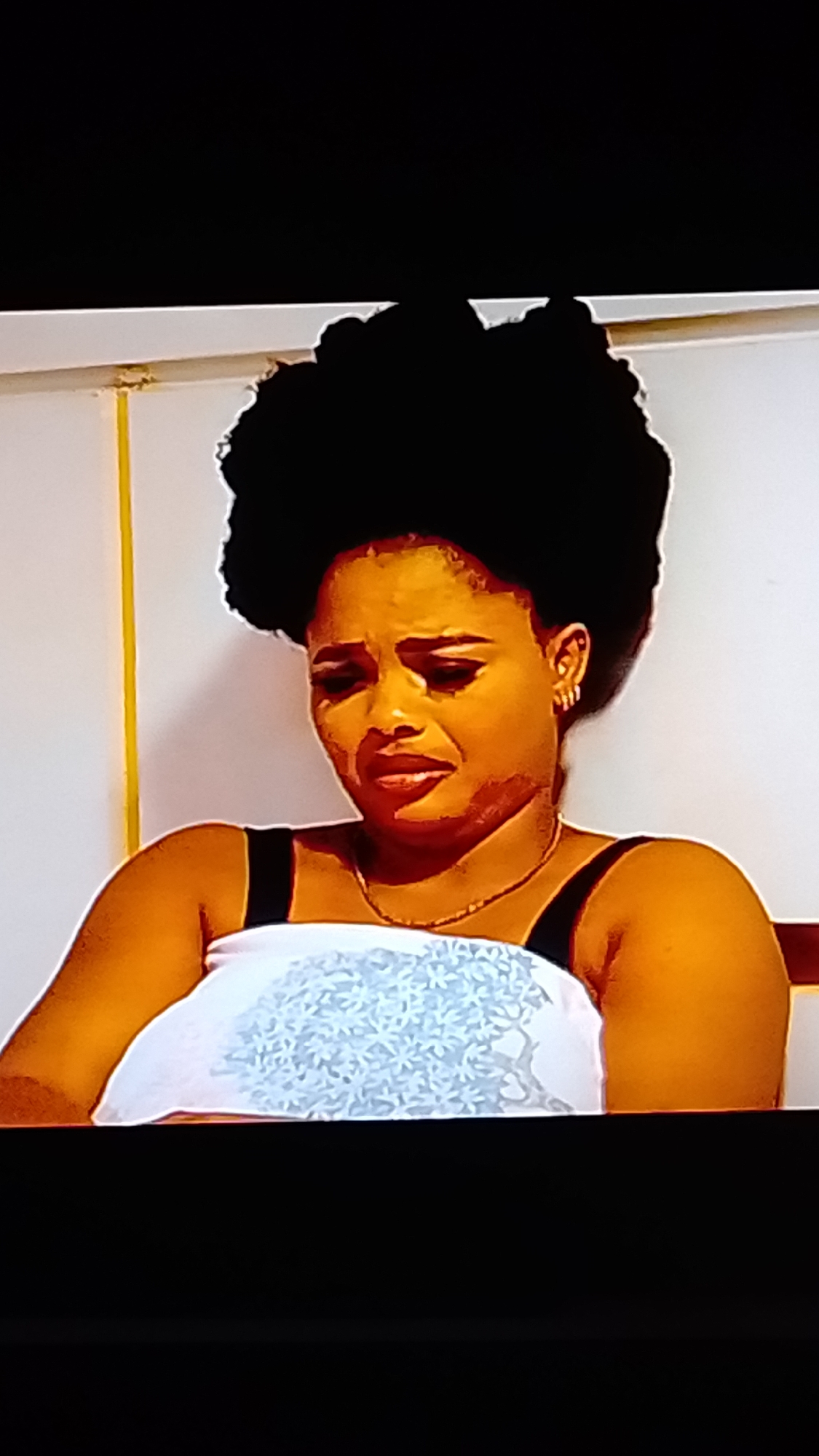 A woman with a large afro hairstyle and dark skin is shown from the chest up. She is wearing a white top with a blue floral pattern and a black strap. She is also wearing a thin gold chain necklace and small hoop earrings. Her face is contorted in a grimace, with her eyes closed and mouth open, suggesting distress or pain. The background is a plain, light-colored wall with a yellow vertical line on the left side. The lighting is warm, casting a golden hue on the woman's skin.