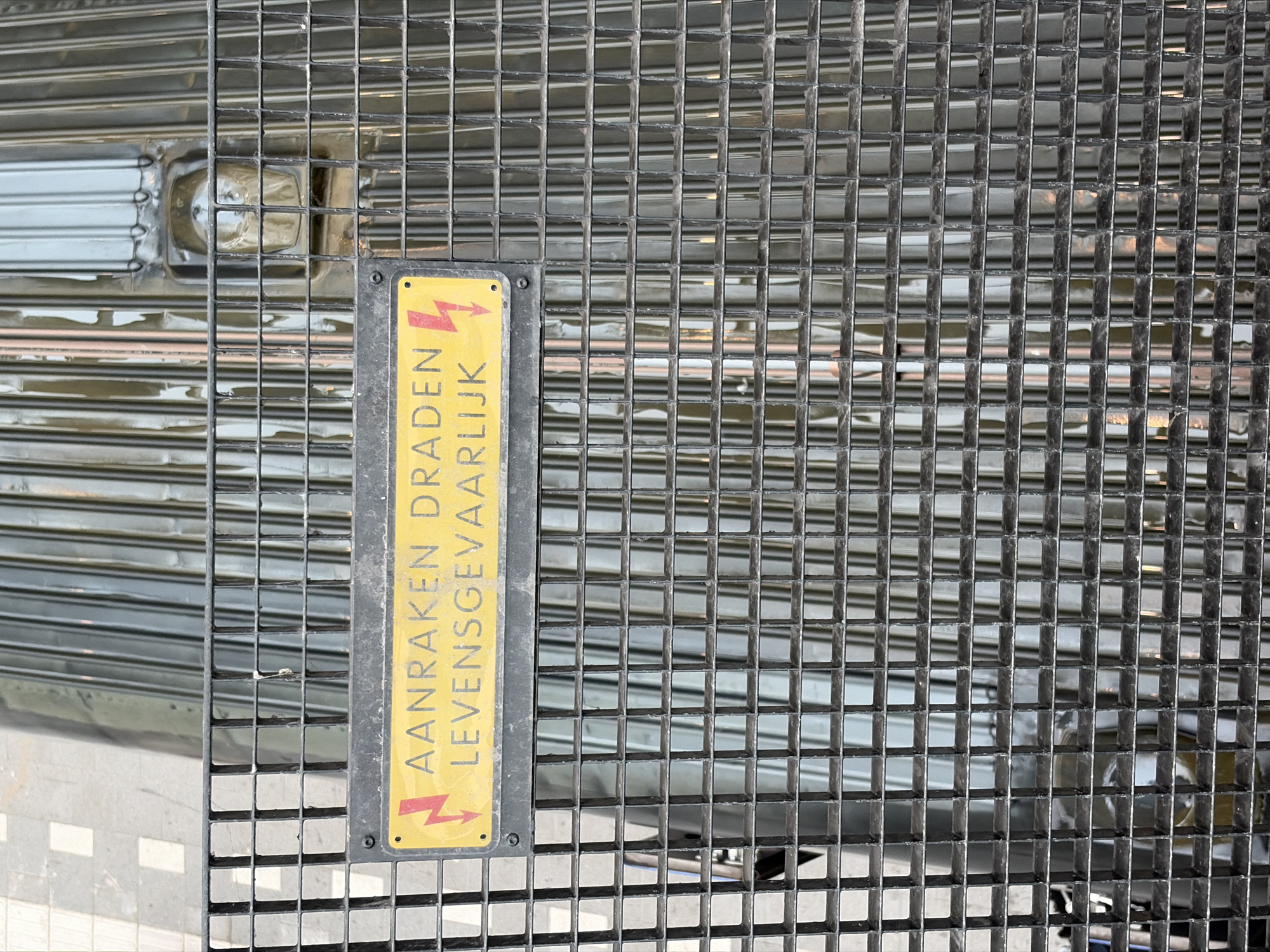 This close-up, eye-level shot captures a dark grey metal grating, likely serving as a floor or cover, in an urban or industrial setting in The Hague, Netherlands. The grating is composed of numerous small, square cells, with some surfaces appearing glossy and reflective, showing blurred reflections of the environment.
A yellow rectangular warning sign is prominently displayed, attached vertically to the grating slightly to the left of the frame's center. The sign features black lettering in Dutch and two red lightning bolt symbols pointing downwards, indicating danger. The text reads: "AANRAKEN DRADEN LEVENSGEVAARLIJK," which translates to "Touching wires is life-threatening." The sign is secured by at least one visible screw or rivet at the top.
Through and beneath the grating, blurred metallic structures are visible in the background, possibly parts of a rolled-up security shutter or similar equipment, suggesting an entrance or access point to a building or utility area. A lighter, textured surface, possibly paving or tiling, is also subtly visible at the bottom left, seen through the grating. The lighting suggests it is daytime, with no specific weather conditions discernible. No human subjects or active events are present; the scene is static, focusing on the warning and the structure itself, emphasizing caution in an area likely involving electrical components or machinery.