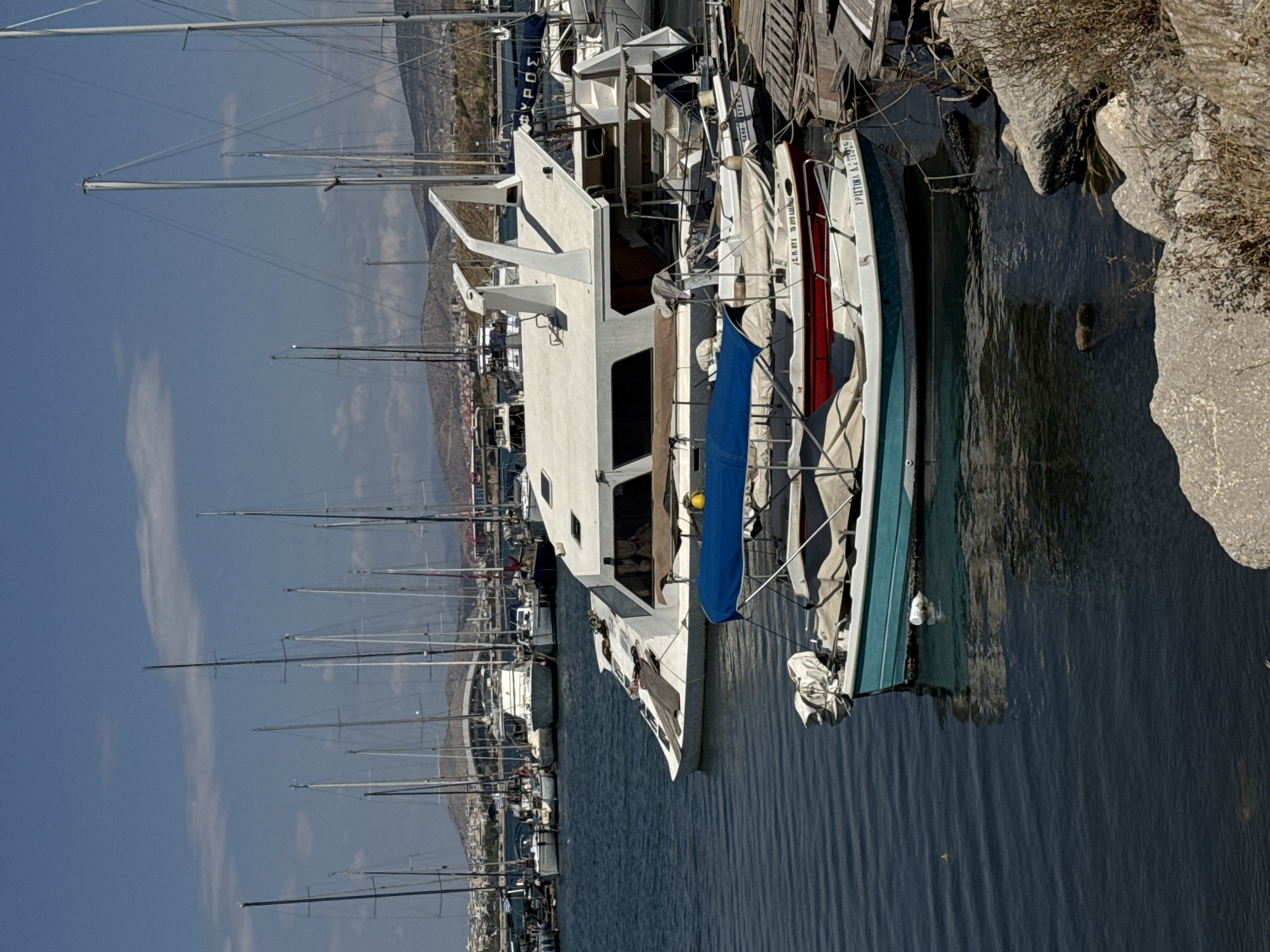 This image depicts a marina in Athens, Greece, likely on a sunny day with clear skies and scattered clouds. The foreground features several boats docked in the dark blue water. The closest boat is a small, teal-colored vessel with a white outboard motor covered by a protective tarp. Behind it, a larger boat with a red hull and a white canopy is visible. Further back, a prominent white multi-hulled yacht with a distinctive angular design and a large canopy structure dominates the mid-ground. Numerous other sailboats with tall masts are moored in the background, creating a dense nautical scene. The shoreline in the distance shows hills with buildings, indicating an urban or semi-urban environment. Rocks and dry vegetation are present along the immediate edge of the water in the foreground. The overall impression is a busy harbor filled with recreational and possibly commercial vessels.