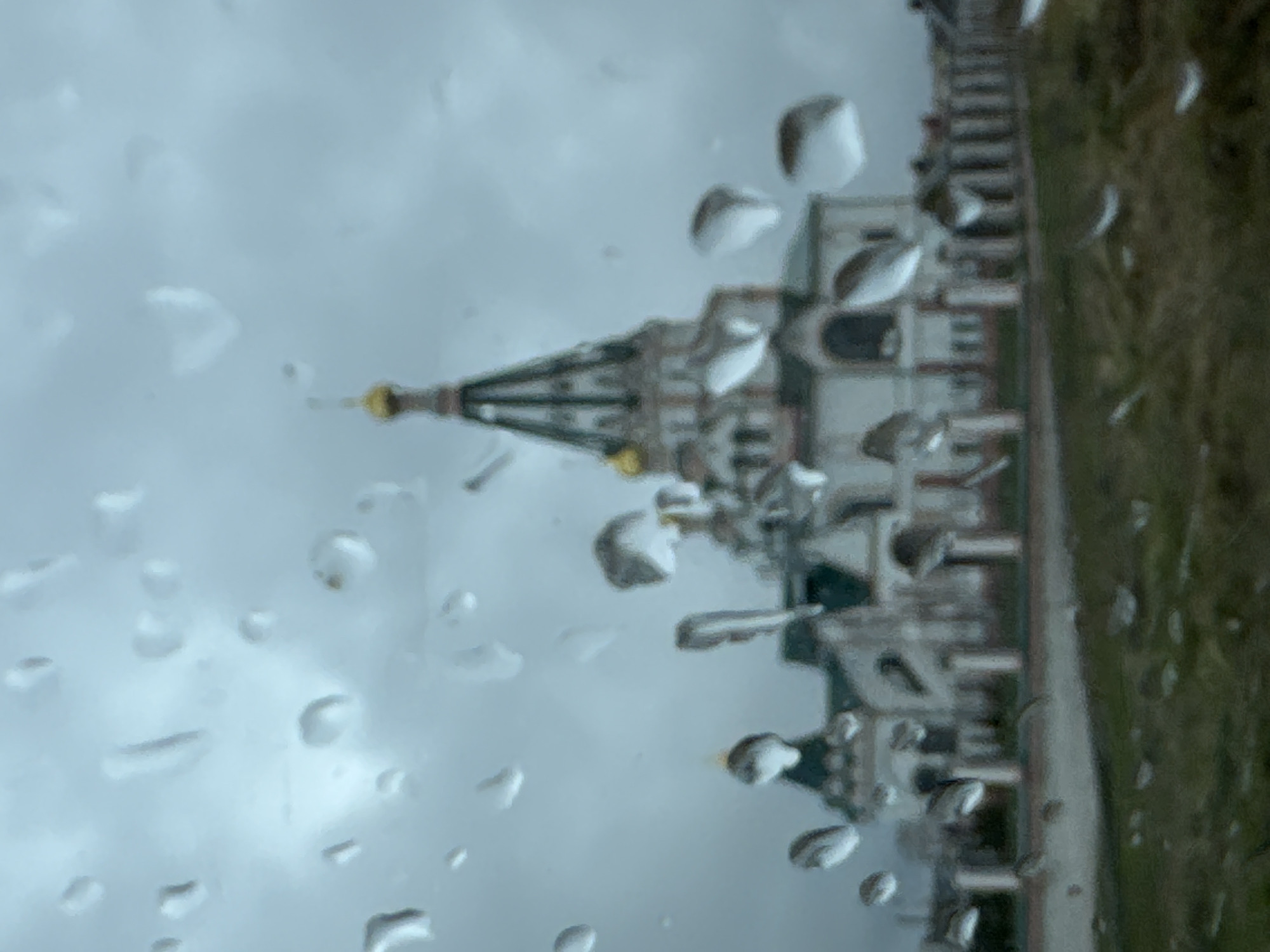 The image depicts a view of the Church of the Holy New Martyrs and Confessors of Russia in Ново-Никольское, Russia, obscured by raindrops on a window. The church's distinctive architecture, including its tower and white exterior with red and green accents, is visible through the blurred view. A cloudy sky is in the background. Part of a sidewalk and greenery are visible to the right of the building.