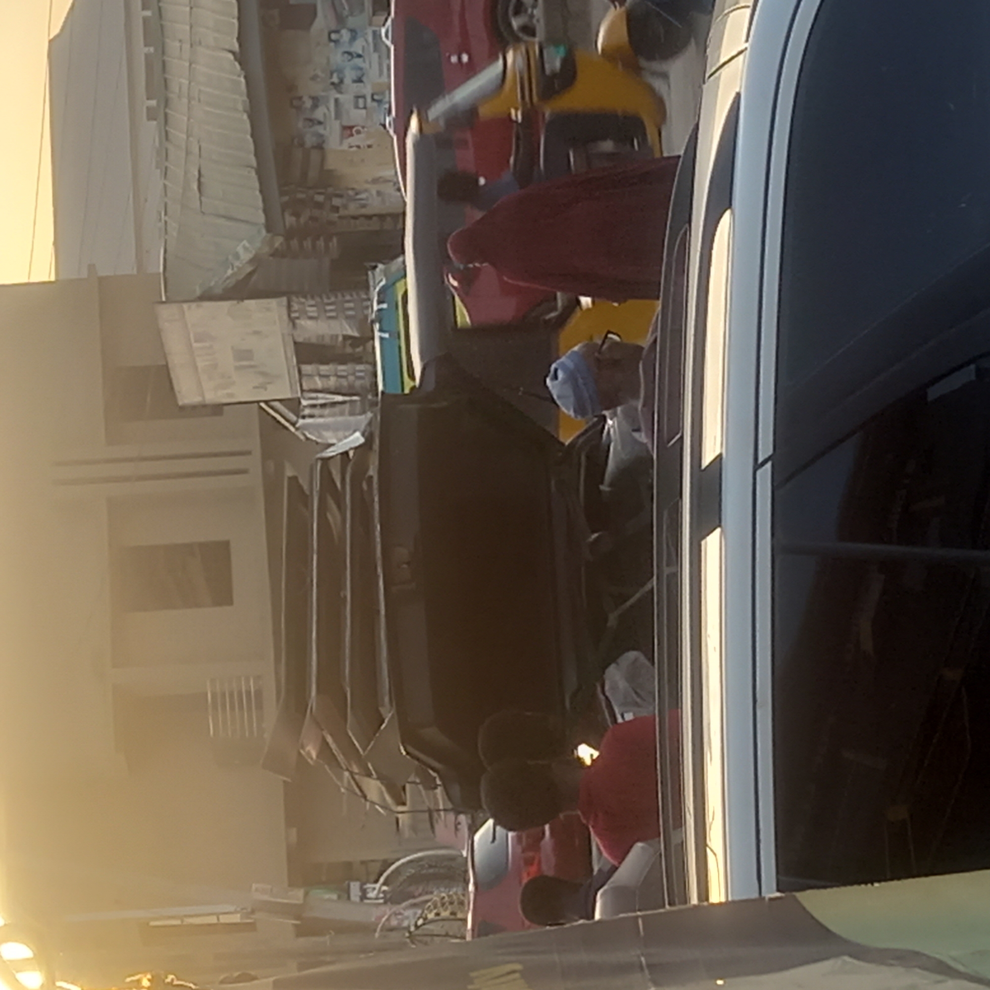 A daytime street scene in Dadi Riba, Nigeria, shows a row of parked cars and vans along a street lined with low buildings and shops. People are visible around the vehicles, some appearing to be engaged in conversations or transactions. A person in a red shirt is sitting, while another person wears a blue head covering. The environment is urban, with visible signs of commercial activity and pedestrians present.