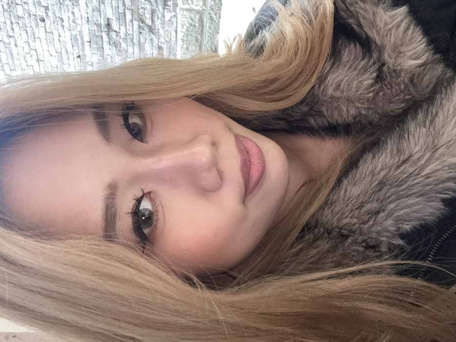 A close-up portrait features a female subject of East Asian appearance, positioned centrally, looking directly into the camera with a slight smile. Her long, light brown hair is parted on her left side and falls over her shoulders. She is wearing a dark-colored jacket with a visible, greyish-brown fur or faux fur collar. Her eyes are dark, possibly grey-brown, with visible eyeliner and eyelashes. Her lips are closed, colored with a light pink or nude shade. The background is out of focus, revealing a light-colored brick or stone wall on the right side, with a horizontal stone or concrete ledge below it. The lighting appears soft and even. The scene is located in Yuzhong District, China.