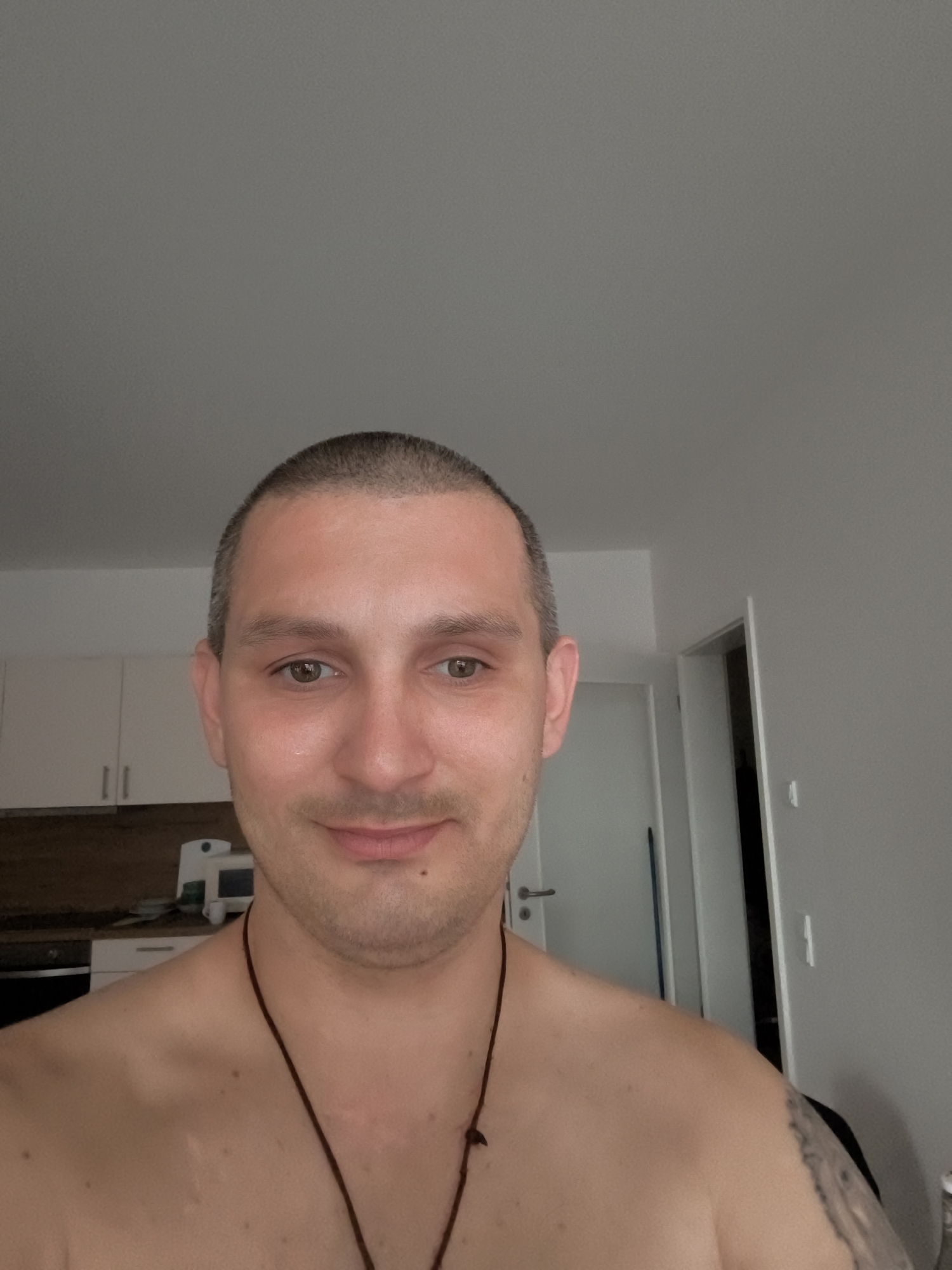 The image is a close-up, self-taken photograph of a fair-skinned, bald man with light brown eyes. He is shirtless and wearing a dark string necklace. He has a faint stubble and a mole below his lower lip. His expression is neutral. In the background, there is a kitchen with white cabinets and a dark countertop, and a doorway leading to another room. The environment appears to be an indoor residential setting. The location of this scene is confirmed as Wolgast, Germany.