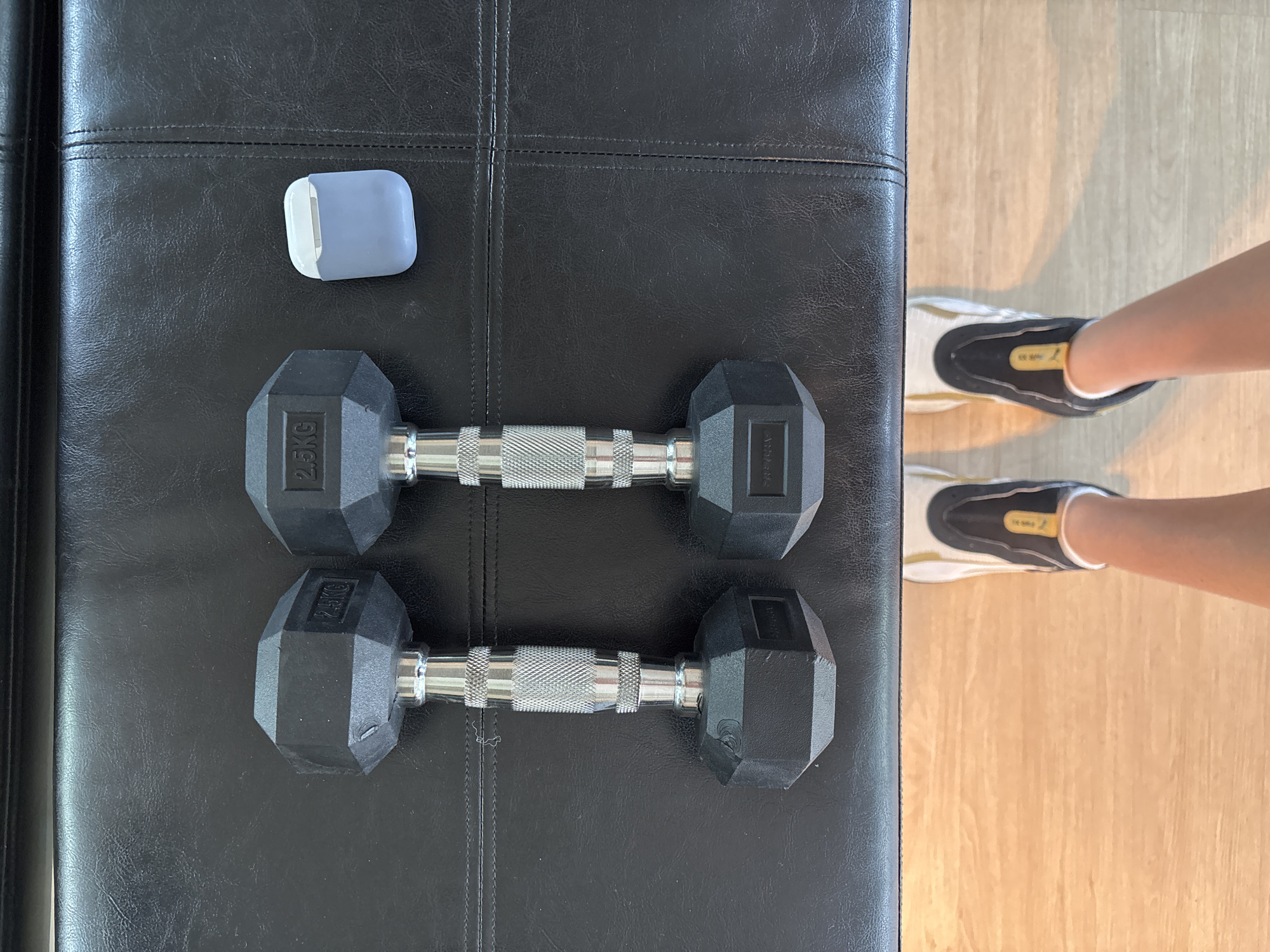 An overhead shot captured in Choeng Thale, Thailand, depicts an indoor scene focused on fitness elements. A substantial black padded surface, resembling a workout bench or sofa, occupies the left and central areas of the frame. On this surface, two black hexagonal dumbbells are arranged horizontally. Each dumbbell has a chrome, knurled handle and prominently displays "2.5KG" on its rubber head, indicating its weight. A light grey and white earbud case, possibly for AirPods, is placed in the top-left corner of the padded surface.
To the right, a light-colored wooden floor is visible. On this floor, the lower legs and feet of a person are seen, clad in black and white sneakers with distinct yellow accents. The person appears to be standing or positioned beside the padded bench. The overall arrangement suggests the person is either preparing for, actively engaged in, or has just completed a light exercise session. The lighting is bright and even, consistent with an indoor daytime setting.
