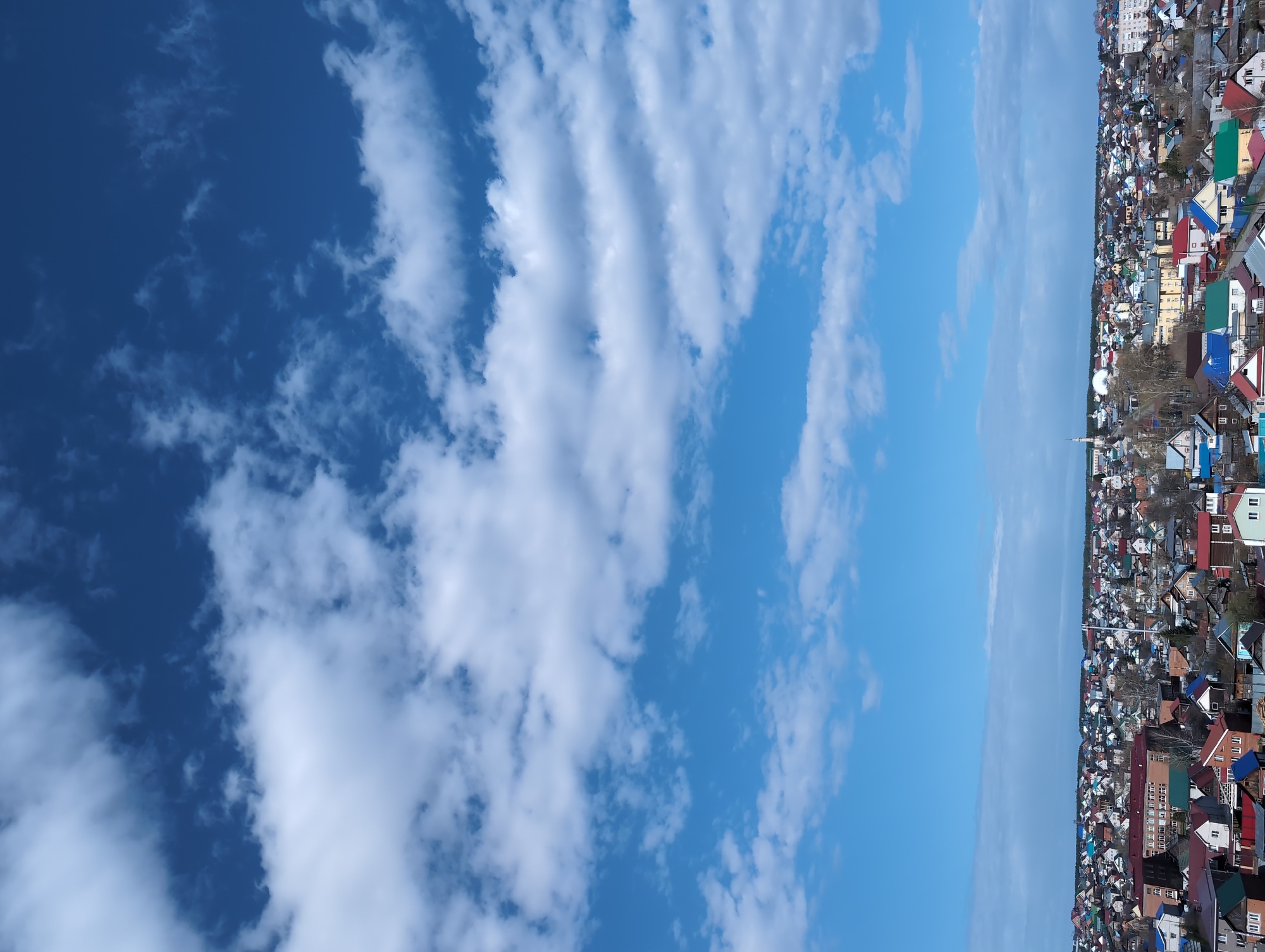 The image presents a wide-angle view of a town or city under a vast sky, appearing to be taken from a slightly elevated position. The sky occupies approximately two-thirds of the frame, showcasing a clear blue expanse with various cloud formations. On the left, denser, more scattered white clouds are visible against a deeper blue, while towards the right, the sky lightens slightly, and a distinct band of elongated, wave-like clouds (possibly undulatus altocumulus or stratocumulus) stretches across the middle.
The lower third of the image depicts a sprawling residential area. Numerous houses and buildings are densely packed, featuring a variety of architectural styles and roof colors, including vibrant reds, blues, greens, and more muted browns and grays. Patches of trees are interspersed among the structures, appearing largely bare, suggesting a season of late winter or early spring. In the distance, a faint, tall, slender structure, possibly a steeple or antenna, rises above the rooftops.
The scene conveys a bright, clear daytime, with good visibility and fair weather conditions. There are no people visible, and no specific activity or event is taking place, presenting a static landscape view. No visible text is present in the image.