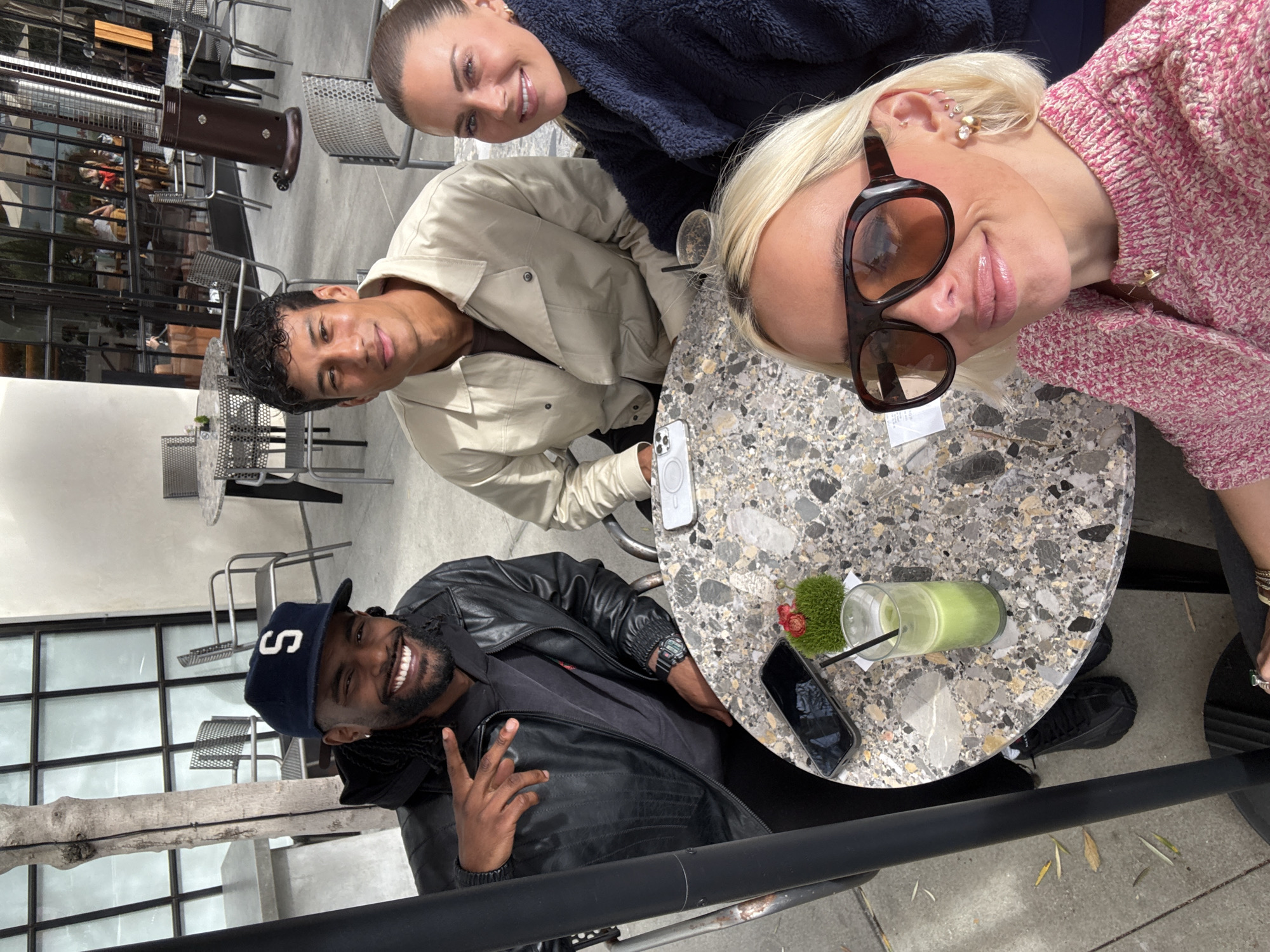 Captured from an elevated perspective, the image features a gathering of four individuals around a round table in an outdoor setting, potentially a café or restaurant patio in Los Angeles, United States of America, inferred from the architectural style and ambiance.
The lower-left quadrant presents a man wearing a black leather jacket over a dark shirt and a navy baseball cap with the letter "S." He smiles and makes a peace sign with his right hand. At the top of the frame, a woman with her hair pulled back is wearing a dark blue sweater. Next to her is a man in a beige jacket. Adjacent to him, to the right side, is a blonde woman wearing large, brown-tinted sunglasses and a pink sweater.
The table, of terrazzo construction, holds various items including a smartphone, a white-cased smartphone, and a glass of green liquid with a straw, adorned with a small decorative plant. The setting includes typical outdoor furniture such as metal chairs and tables, situated on a paved surface. The background shows glass-paneled walls with reflections, indicating buildings or structures nearby. The overall lighting suggests it's either late afternoon or early morning.