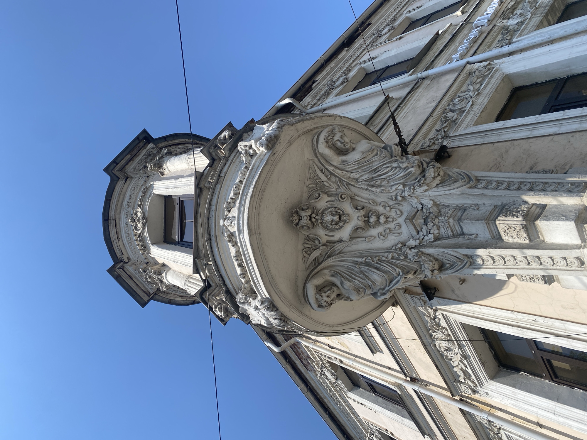 A low-angle shot captures a corner of a light-colored building against a clear blue sky. The building features intricate architectural details, including a circular balcony supported by sculptures of reclining figures. Ornate carvings adorn the window frames and cornices. Electrical wires run diagonally across the upper left corner of the frame. The photograph's location is Заволжье, Russia.