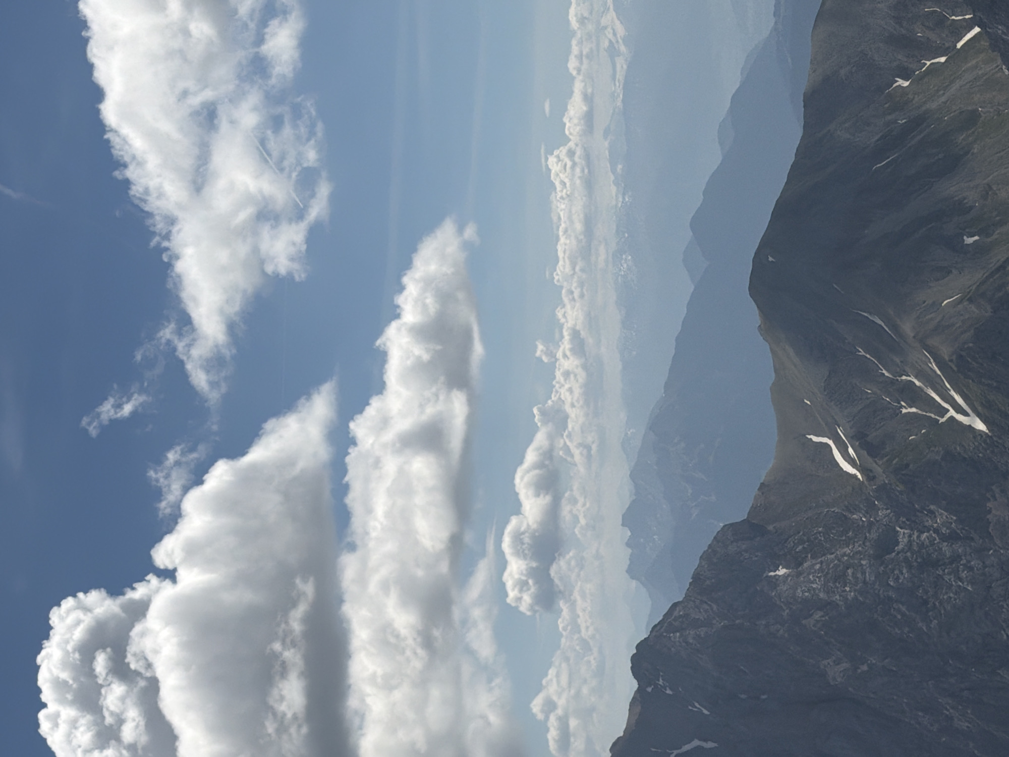 The image displays a panoramic view of a mountain range under a partly cloudy sky. The foreground features a dark, rugged mountain peak with patches of snow. In the middle ground, successive layers of mountains recede into the distance, becoming progressively lighter and hazier due to atmospheric perspective. The sky is a bright blue, dotted with large, fluffy white cumulus clouds. There are no people or man-made structures visible in the scene. This mountainous landscape is characteristic of the Bavarian Alps, an area that includes Garmisch-Partenkirchen, Germany.