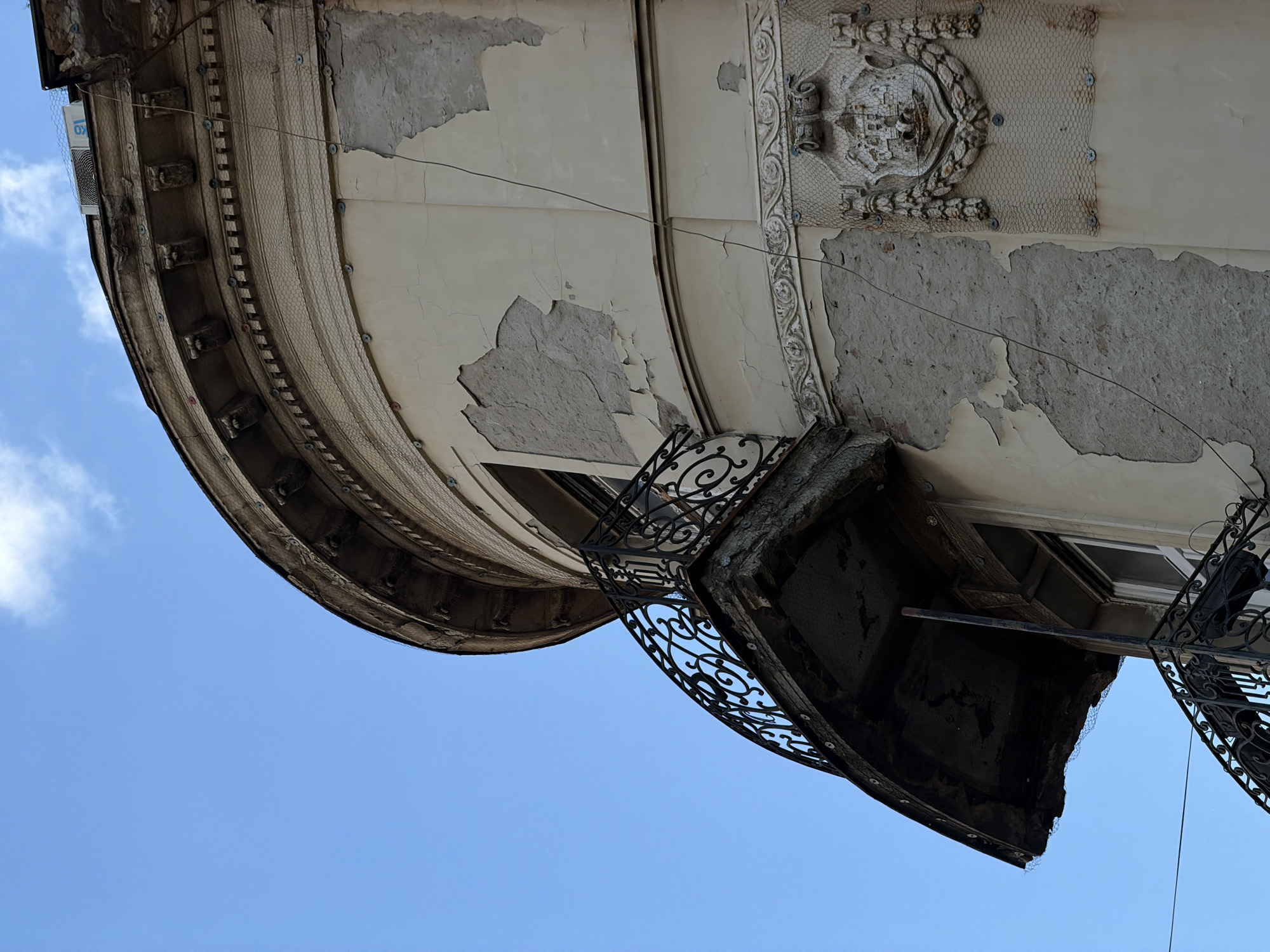 An upward-angled shot shows a section of an old building in Belgrade, Serbia, exhibiting signs of disrepair. The lower portion of the frame is filled with a clear blue sky with a few scattered clouds. The building's facade is peeling and crumbling in several areas, revealing the underlying concrete structure. A wrought-iron balcony juts out from the building's curved surface. The balcony is blackened and weathered, suggesting neglect and age. The building's architectural details include decorative moldings and a crest or shield-like ornament, also partially damaged and covered with protective netting. An air conditioning unit is visible on the left side of the building. Electrical wiring is exposed along the building's facade. The image emphasizes the building's decay.