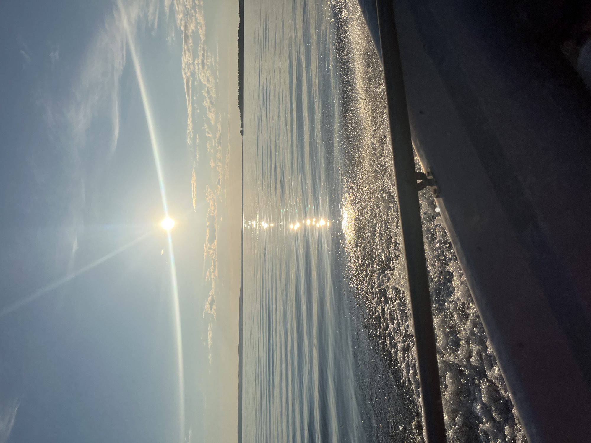 The image captures a serene scene from a boat moving across a body of water, likely in Siukterka, Russia. The sun, positioned high in the sky and casting a brilliant glare, suggests it is midday. Wispy clouds streak across a clear blue sky. The water's surface is calm, with gentle ripples creating a textured pattern. Sunlight reflects off the water, forming shimmering lines that stretch towards the horizon. The distant shoreline is visible as a dark, silhouetted line, hinting at land or a forest. The right side of the frame is dominated by the side of the boat, with frothy white water churning in its wake, indicating movement. The overall mood is peaceful and tranquil, evoking a sense of calm and the beauty of nature on a sunny day. There is no visible text in the image.