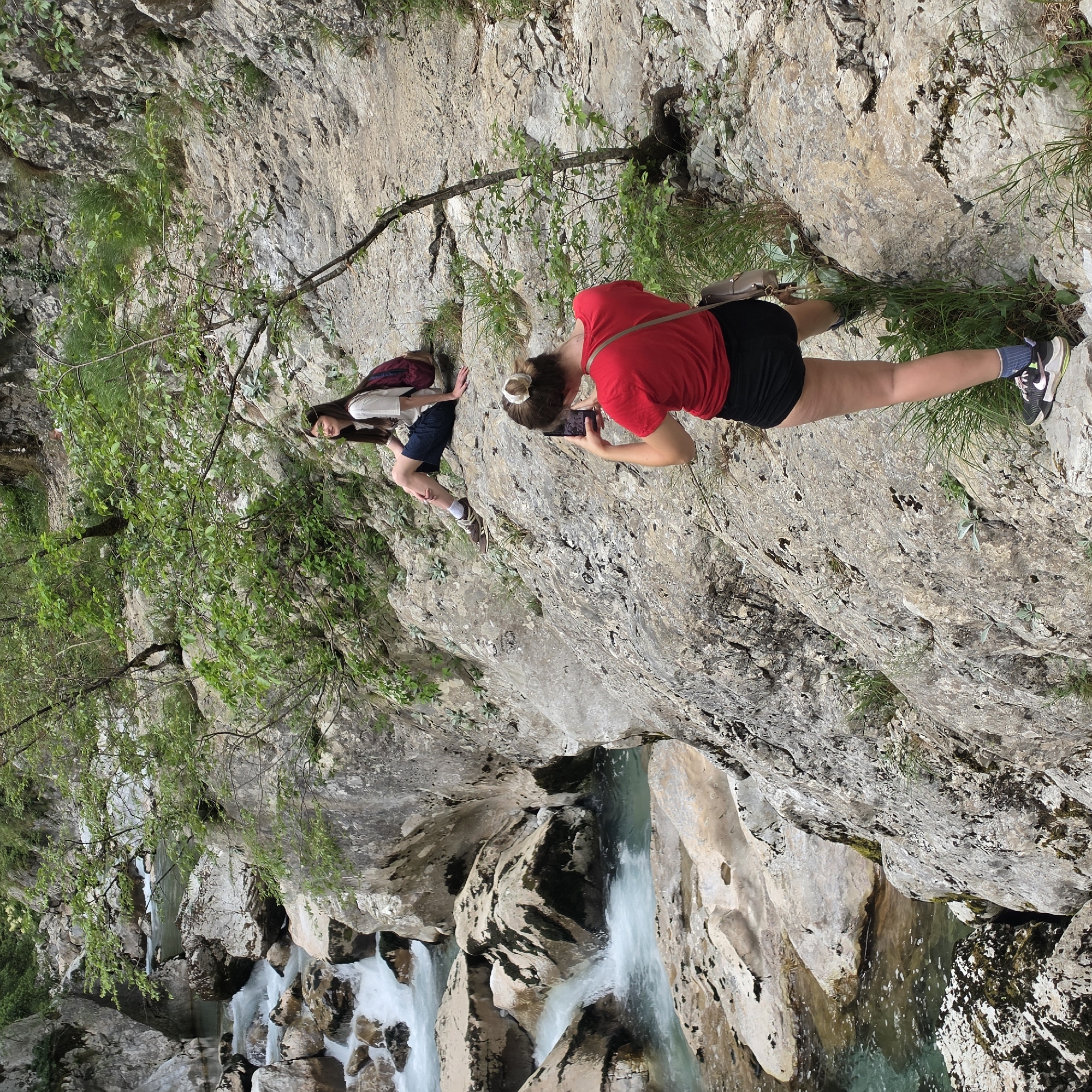 Two young women are perched on a rocky ledge overlooking a river. One woman, wearing a red shirt and black shorts, is taking a photo with her phone. The other woman, wearing a white shirt, is next to her. The river below flows through a rocky gorge. Green vegetation is visible on the rocks. The location is identified as Lazba, Montenegro.
