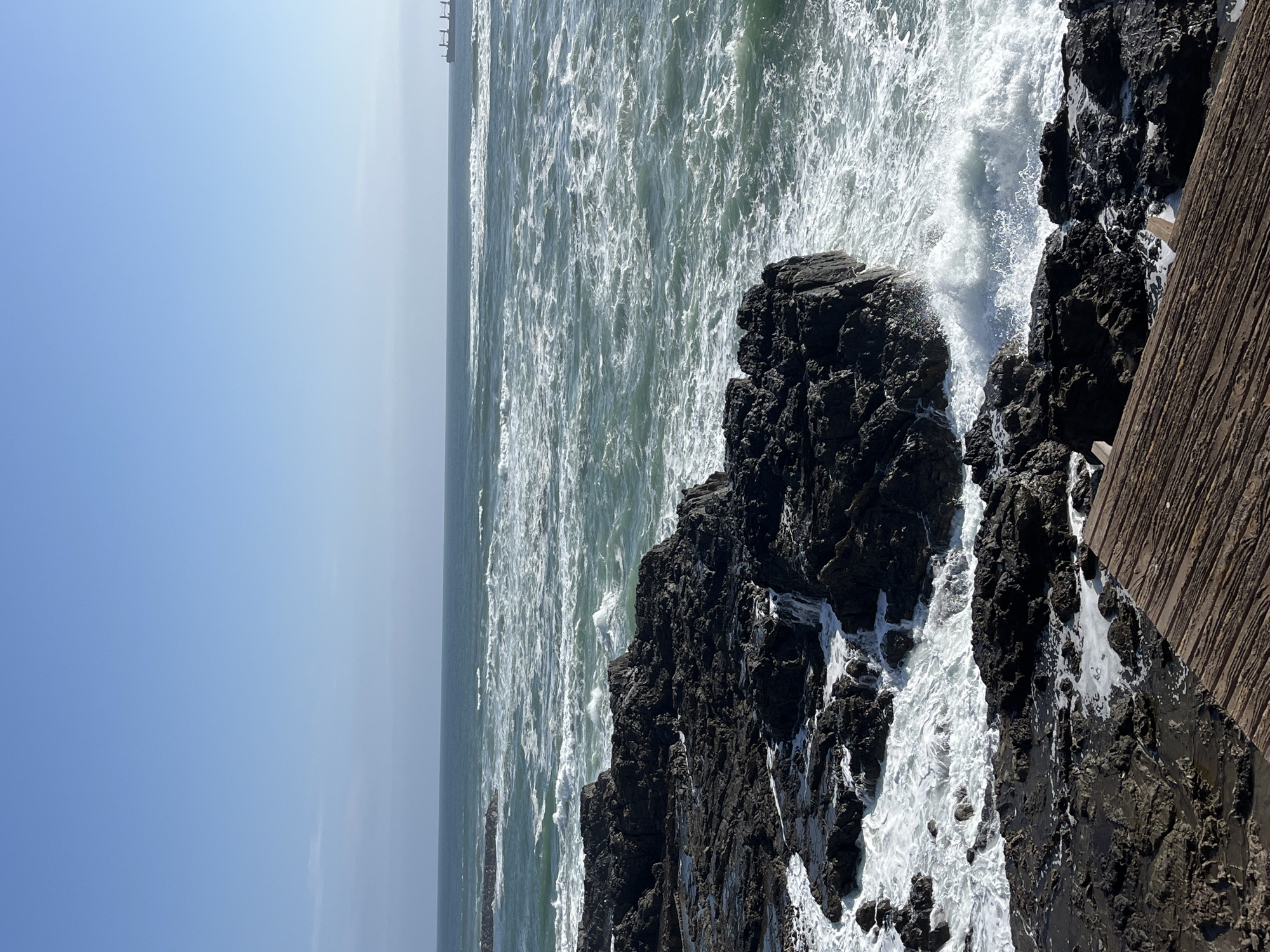 The image depicts a coastline in Arica, Chile. The ocean is agitated, with white foamy waves crashing against dark, jagged rocks. A wooden barrier runs along the right edge of the frame, suggesting a walkway or cliff edge. In the distance, a pier structure is faintly visible on the horizon. The sky is clear and pale blue.
