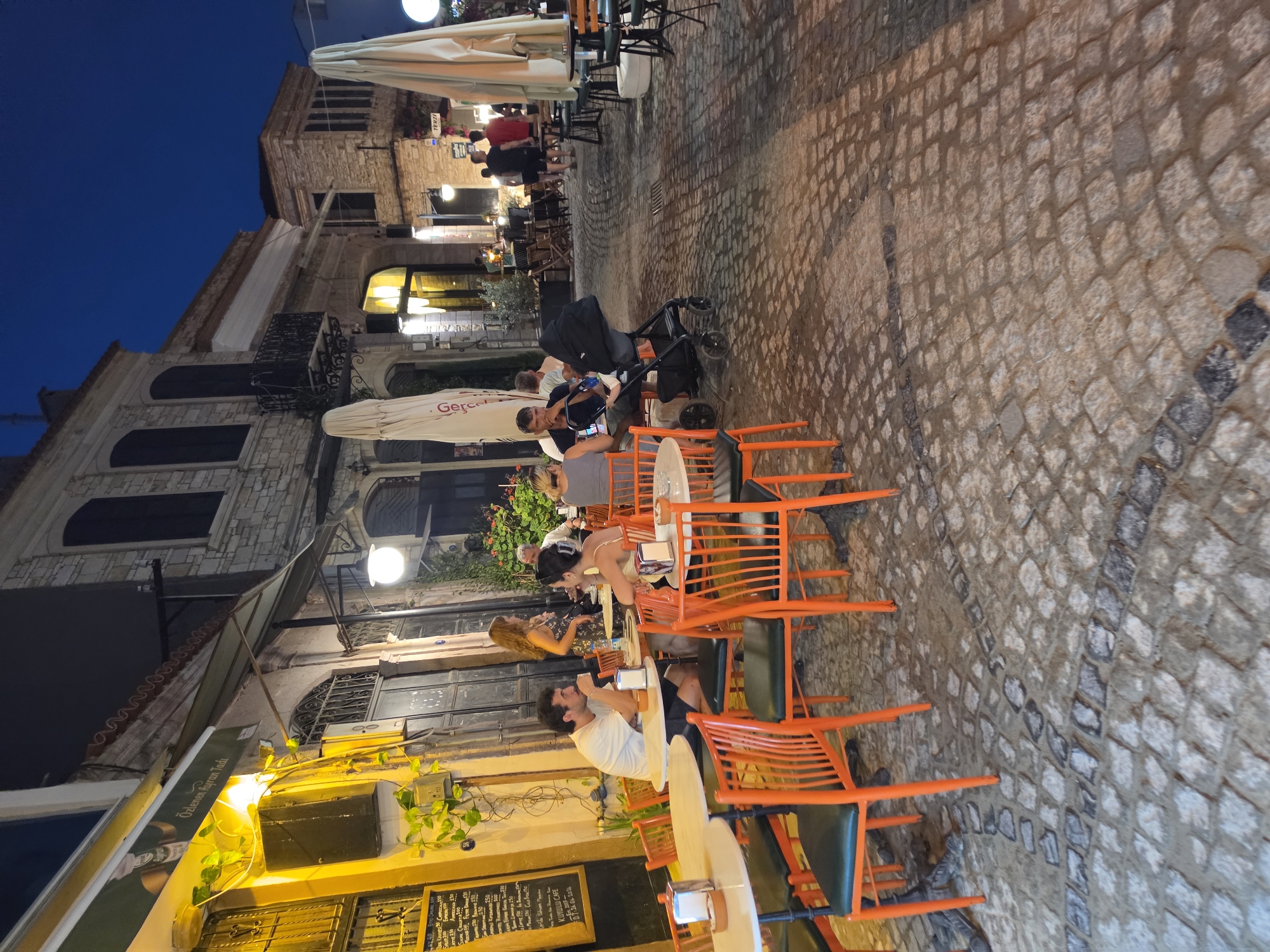 The image depicts an outdoor seating area of a cafe or restaurant at dusk in the city of Urla, Turkey. The ground is paved with cobblestones. Several tables with orange chairs are set up. In the foreground, a group of people are seated at a table. A man in a white t-shirt is looking forward, while a woman in a yellow top is facing him. To their right, a baby stroller is parked near another table where a man is sitting. In the background, more people are walking along the street and seated at tables. The buildings lining the street are made of stone and have arched windows and balconies. Outdoor umbrellas are visible, and string lights illuminate the area. A menu is posted on a wall on the left side of the image. The sky is a deep blue, indicating the transition from day to night.