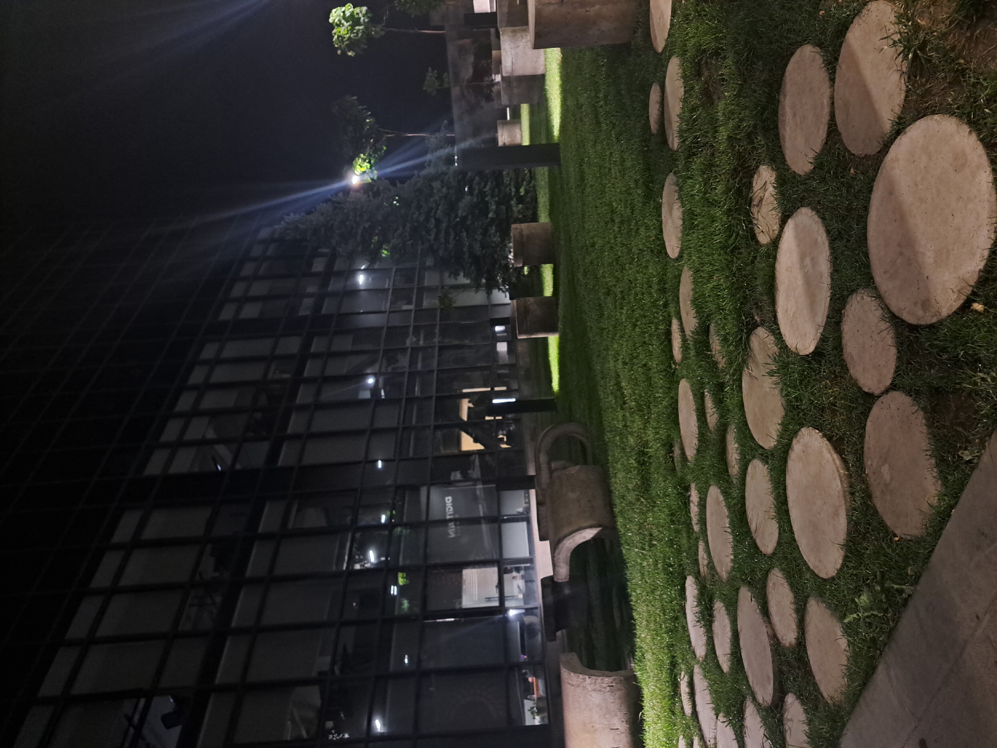 Here is a description of the image:
The image shows an outdoor space at night, likely in Yerevan, Armenia. A modern building with a grid-like window facade is on the left side. A green grassy area runs parallel to the building, featuring a winding path made of large, circular concrete stepping stones. Scattered throughout the grass are short, cylindrical concrete pillars or blocks, some appearing to hold greenery on top. The scene is lit with soft lighting, creating a relaxed atmosphere. The image is taken from a slightly low angle, enhancing the sense of depth.