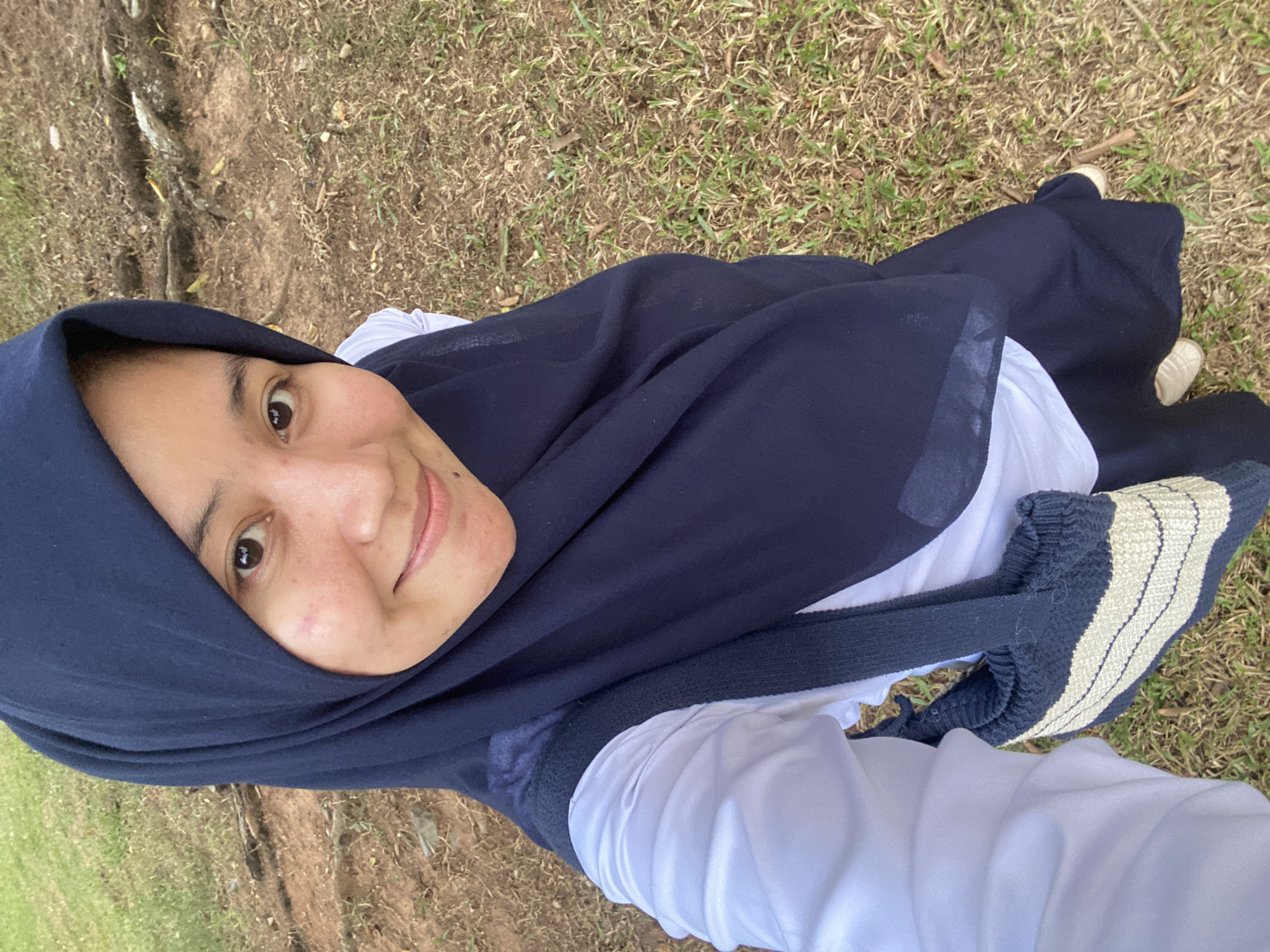 A young woman wearing a navy blue hijab and a white shirt with a dark blue vest is taking a selfie outdoors. She is smiling and looking at the camera. Her left shoulder is visible, and she is holding a navy and cream striped bag over her left shoulder. The background consists of dry grass, soil, and tree roots, suggesting an outdoor, possibly park-like setting. The lighting appears natural, indicating daylight. The image is taken from a slightly high angle, looking down at the woman. There are no other people or distinctive landmarks visible in the image. The environment suggests a natural, unpaved area, potentially a public park or a less manicured outdoor space. There is no specific indication of the city of Banjarbaru, Indonesia, within the image itself.