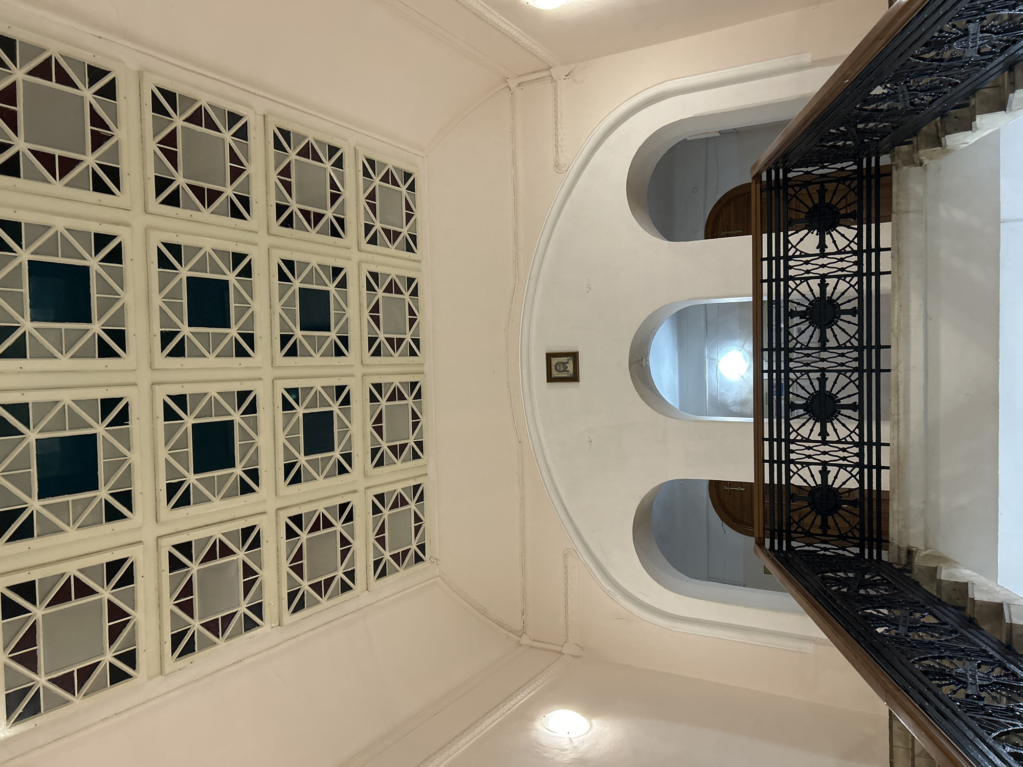 The image shows an interior view of a staircase and hallway. The ceiling features a decorative pattern of square panels, each containing a geometric design with triangles in shades of gray, dark blue, and maroon, surrounding a central square. The walls are painted a pale pink. Above the arched openings of the hallway, a framed religious icon is visible. A wrought-iron railing with circular decorative motifs lines the staircase, which appears to be made of stone or marble steps. The hallway has arched doorways, one of which reveals a wooden door with a cross. The scene is likely located in Tomsk, Russia, given the presence of the religious icon and the architectural style, which is consistent with historical buildings in the city. There are no people visible in the image.