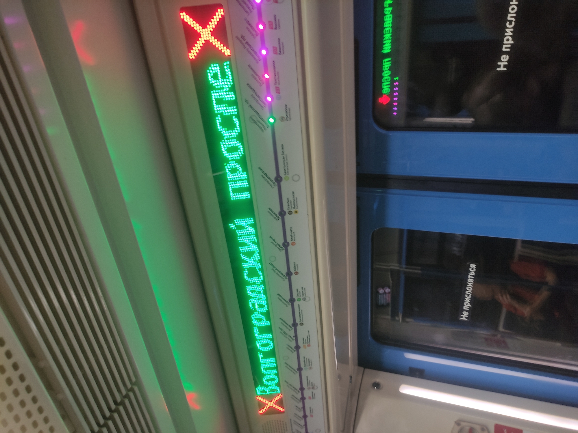 Here's a forensic description of the image:
**Scene**: Interior of a Moscow Metro train car.
**Subject**: An illuminated train route map and signage next to the door.
**Details**:
* The route map uses LED displays to show the current station ("Volgogradsky Prospekt") with green lettering. The prior and future stations along the Tagansko-Krasnopresnenskaya line (Line 7, indicated with purple lights) are displayed.
* A red 'X' marks the current location. The name of the line is displayed in reverse on another lit display.
* Adjacent to the illuminated map is a train door with reflective surfaces. A sign in Russian is displayed on the window, which translates to "Do not lean".
* The train car interior has ribbed paneling.