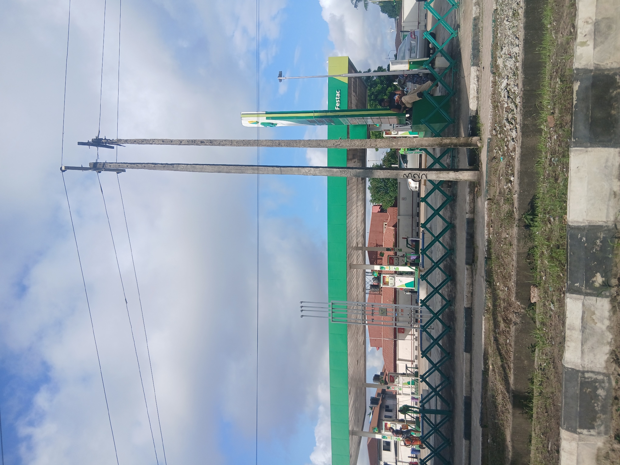 This is a wide-angle, eye-level, daylight photograph of a Festac Gas Station in Isunba, Nigeria. The scene is dominated by the bright green canopy of the gas station, supported by white pillars, stretching across the frame. Several fuel pumps are visible under the canopy, some with green and white branding. A tall, green and white sign with the "Festac" logo stands to the right.
In the foreground, a green, retractable barrier with a diamond pattern divides the street from the gas station forecourt. A man in a dark shirt and light-colored pants is sitting on the green barrier, facing towards the gas pumps. To the left of the scene, two individuals in work uniforms are attending to something near the fuel pumps. A silver car is parked to the left of the forecourt, and another silver car is visible further back, near the entrance to the station.
Several electrical poles with wires crisscrossing the sky are prominent in the upper half of the image, with one pole appearing to be damaged or leaning. The sky is partly cloudy, with patches of blue visible between white cumulus clouds. The ground in front of the barrier is paved with concrete and has patches of dry grass and debris. Beyond the gas station, residential buildings with red-tiled roofs are visible.