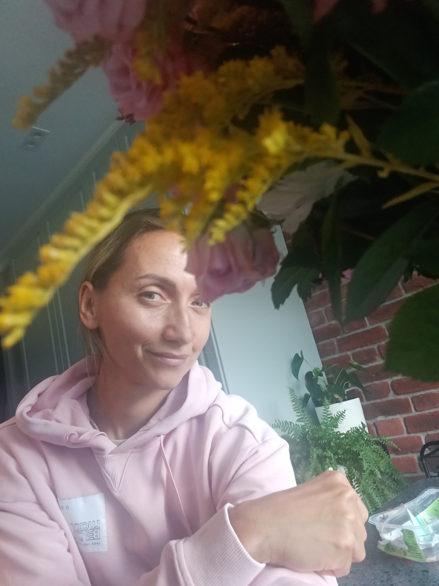A woman with blonde hair pulled back is in the foreground, smiling at the camera. She is wearing a light pink hooded sweatshirt with a white patch on the left chest that has black text on it. A bouquet of flowers, including pink roses and yellow sprigs, is partially obscuring the view of the woman. The background shows a wall of red bricks and a white pot with green plants. The scene appears to be indoors, possibly in a home or a cafe, in the city of Sampo (Krasny Posyolok, Russia).