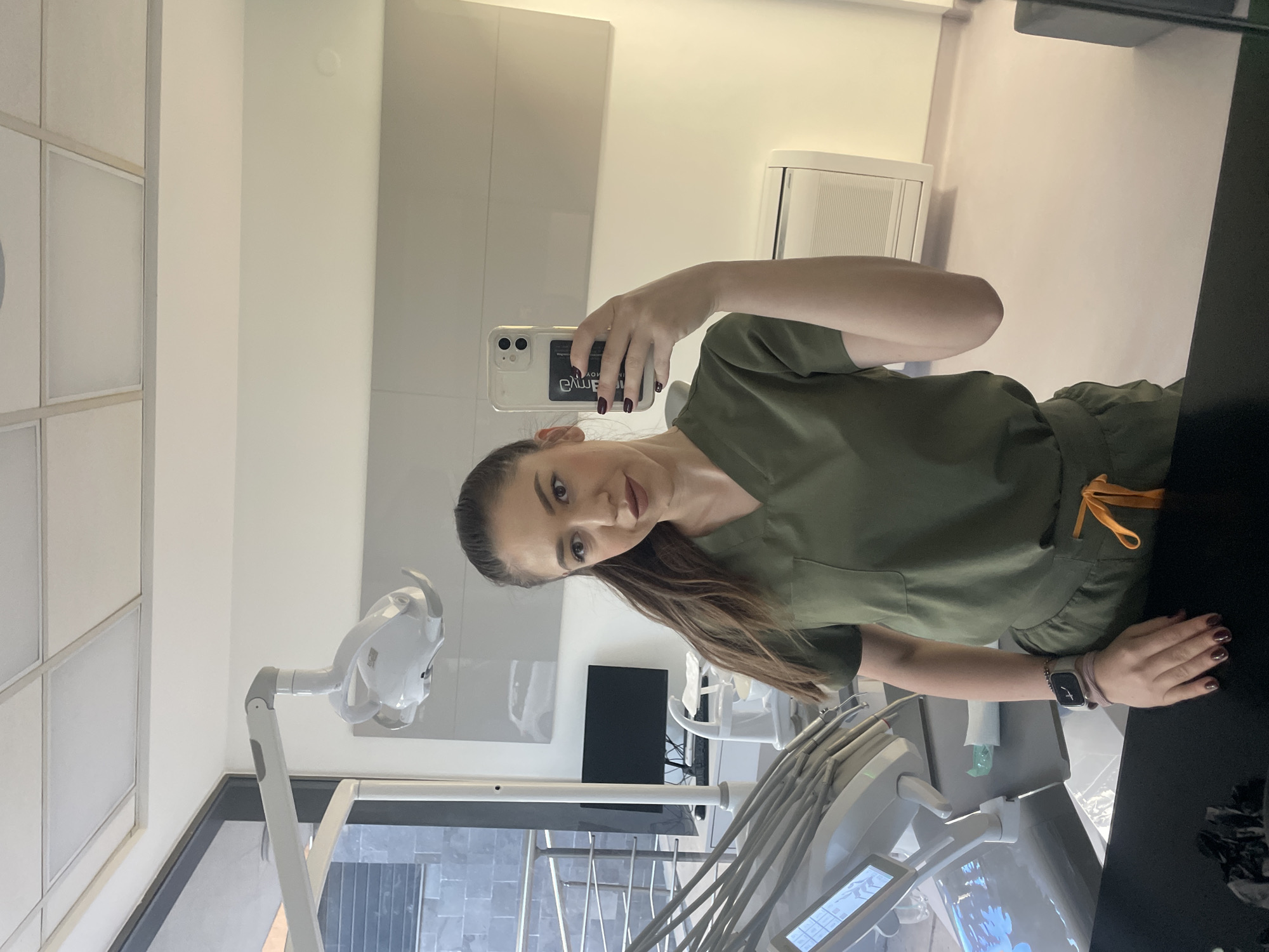 The photograph depicts a woman in green medical scrubs holding a smartphone and taking a selfie. She has dark hair, and is wearing a wrist watch. The setting appears to be a dental office, indicated by the presence of a dental chair, associated equipment, and cabinetry. The background suggests a modern or recently renovated interior, with white and gray color schemes. Based on the smartphone, the city of Sofia, Bulgaria is not evident in the image. The scene appears to be sterile and professional.