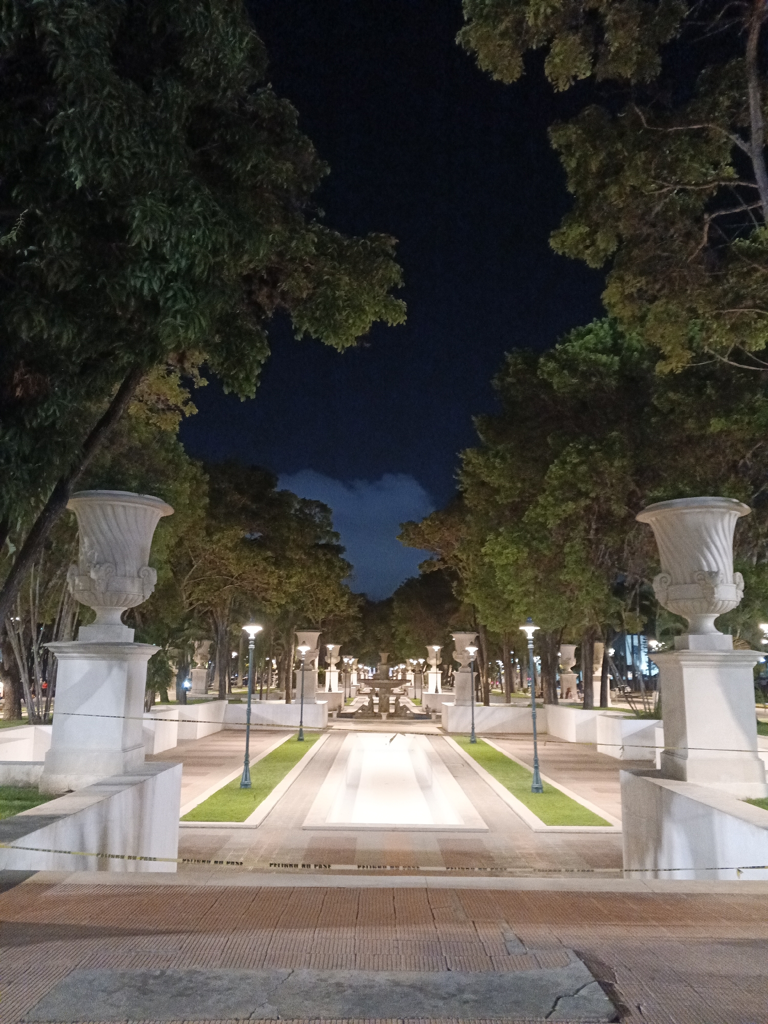 The image depicts a symmetrical pathway in a park setting at night. Tall trees line both sides of the pathway, with lush green foliage visible against a dark blue night sky. The pathway is paved with brick-like tiles in the foreground and transitions to a lighter-colored, smooth surface further into the park. Along the sides of the pathway, there are low white walls, some topped with ornate white vases. White lampposts with glowing orange lights are spaced evenly along the pathway, illuminating the scene. In the distance, a fountain or water feature is visible, also illuminated. The overall impression is of a well-maintained, formal park area designed for evening strolls. The location is identified as Parque Los Caobos in Caracas, Venezuela. There are no people visible in the image. Yellow caution tape with the text "PELIGRO NO PASE" is present across the foreground, indicating a restricted area.