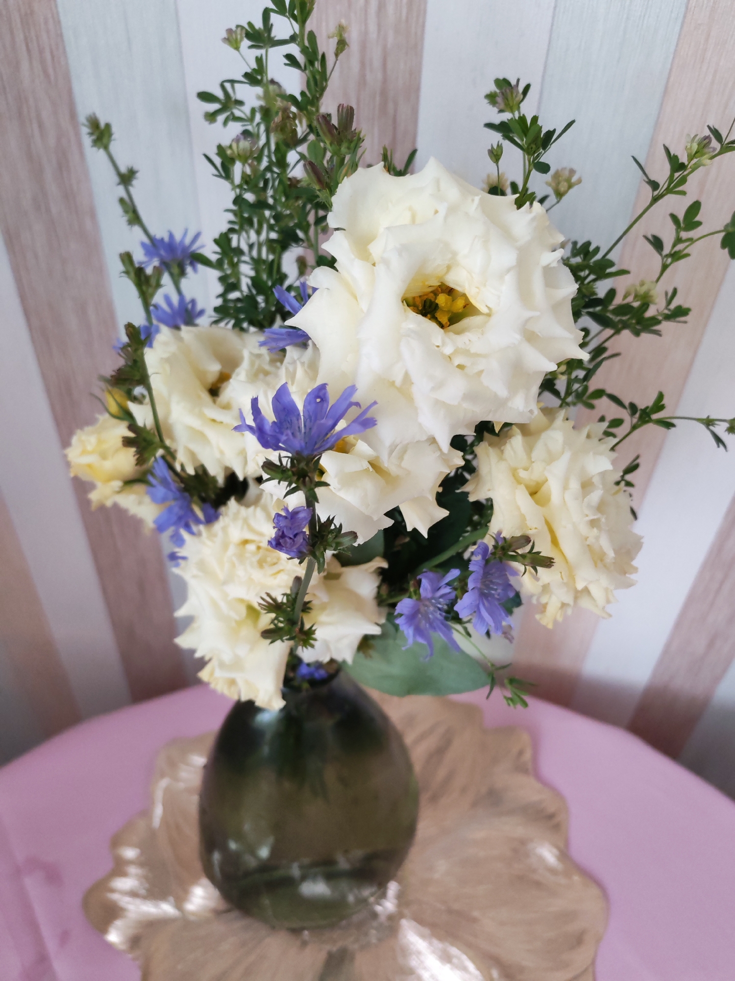 A close-up, eye-level, vertical shot captures a bouquet of cream-colored Eustoma flowers and small, blue Chicory flowers in a dark green glass vase. The vase rests on a golden, decorative placemat with a petal-like edge, which in turn sits on a pink tablecloth. In the background, a wall is decorated with alternating light wood and pale pink vertical stripes. The scene appears to be indoors, possibly in a home in Izhevsk, Russia, given the context.