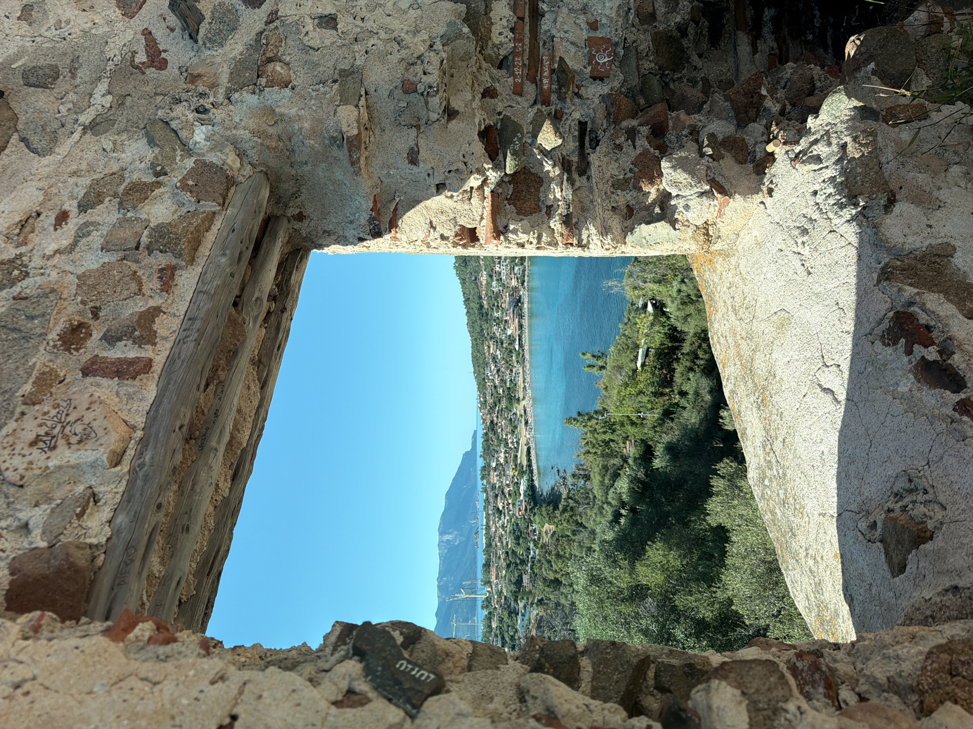 The image is a view through a stone window opening in Tortolì, Italy, revealing a coastal town nestled between lush green hills and a vibrant blue sea. In the foreground, the weathered stone walls of the structure frame the scene, with visible bricks and mortar. A wooden beam spans the top of the opening. Beyond the window, the town is densely populated with buildings, many featuring terracotta roofs. A bay is visible with clear turquoise water, dotted with small boats. In the distance, mountains rise, their peaks partially obscured by haze. The sky is a clear, bright blue. Several construction cranes are visible in the background near the mountains. Vegetation, including olive trees, covers the hillsides. The overall impression is one of a picturesque Mediterranean landscape bathed in bright sunlight.