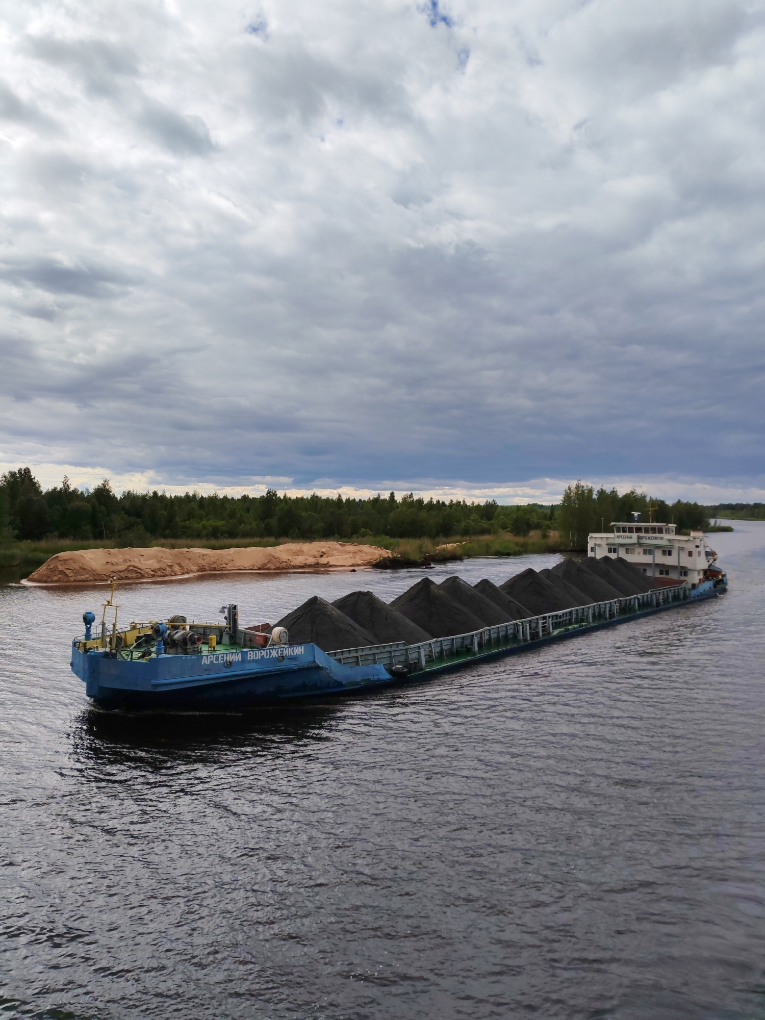 The image shows a blue barge filled with dark gray cargo, possibly coal or gravel, navigating a wide, calm river. The barge has the name "АРСЕНИЙ ВОРОЖЕЙКИН" written on its side in Cyrillic script. Behind the barge, a sandy embankment is visible on the left, topped with trees. A dense green forest lines the far bank of the river. The sky is overcast with dramatic gray clouds. The scene appears to be located in or near Ozernoe Ust'e, Russia, given the context of the provided location. The water is dark and reflects the cloudy sky. There are no people clearly visible in the image. The overall impression is one of industrial activity within a natural landscape.