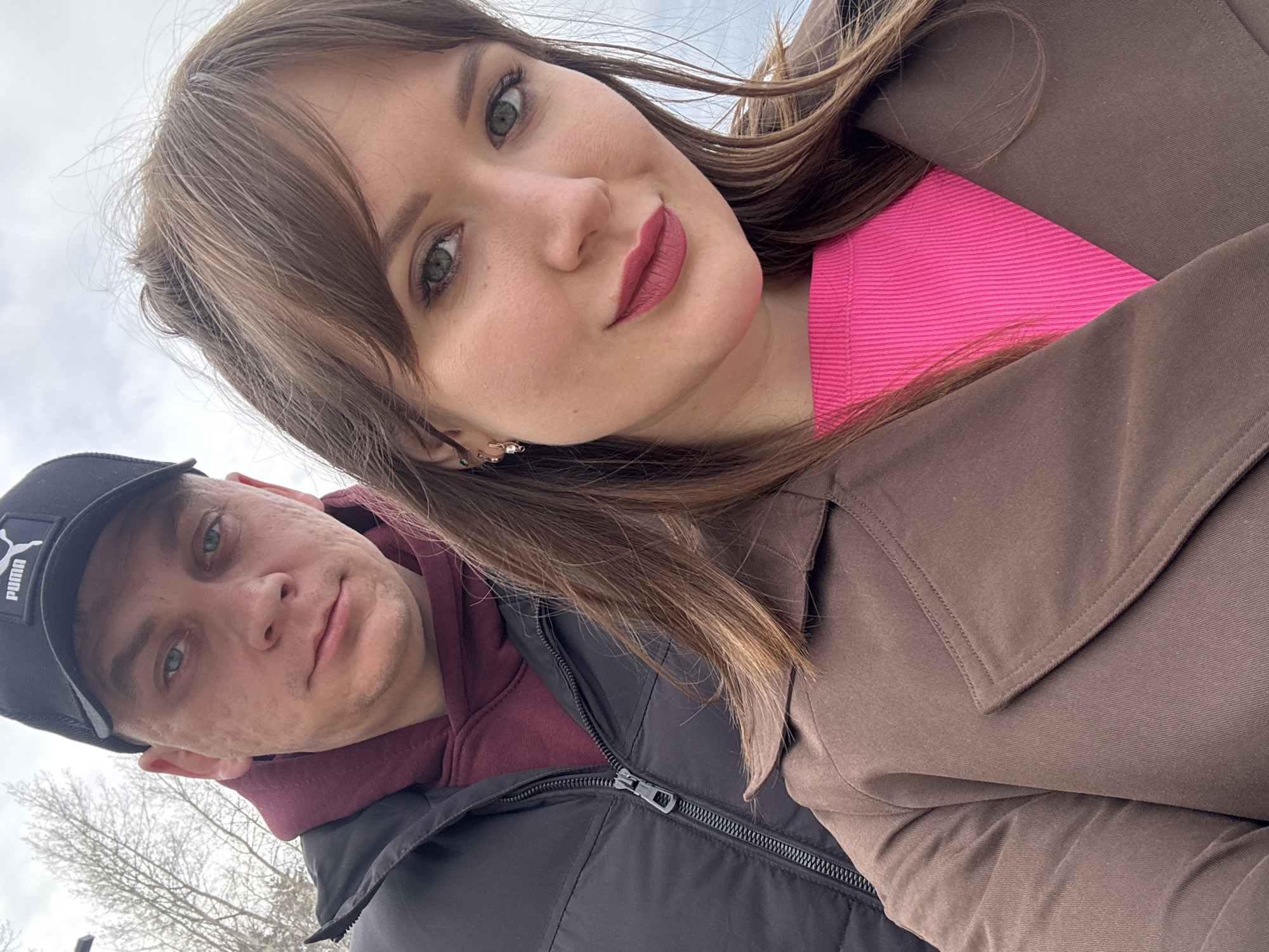 This close-up, tilted shot captures two individuals, a woman and a man, likely posing for a selfie outdoors in Chaikovsky, Russia. The woman is positioned prominently on the right side of the frame, looking towards the camera with a subtle smile. She has long, light brown hair, green eyes, and is wearing pink/mauve lipstick. Her attire includes a bright pink top or lining visible beneath a brown, collared coat, and she wears an earring in her visible ear.
The man is situated slightly behind and to the left of the woman, also looking towards the camera with a neutral expression. He is wearing a black baseball cap with the white "PUMA" logo clearly visible, a maroon hooded sweatshirt, and a black puffer jacket with a distinct zipper.
The background is softly blurred but shows an overcast, light-grey sky and the bare branches of trees, indicating an outdoor setting, possibly a park or residential area. The warm clothing worn by both subjects suggests colder weather, consistent with late autumn, winter, or early spring. The diffused light from the cloudy sky indicates it is daytime.
