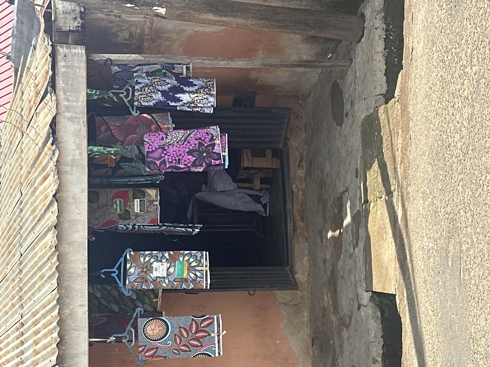 The image depicts the exterior of a rustic shop or stall in the city of Unknown, Unknown, under bright daylight casting strong shadows. The structure features a weathered, corrugated metal roof that extends forward, supported by a horizontal beam. The walls are a textured, reddish-brown, possibly plaster or mud, showing signs of wear and a rough concrete base.
The main subject is a display of patterned fabrics hanging from various clothes hangers (some light blue plastic, some dark metal) under the roof overhang, suggesting the shop sells textiles.
- On the far left, a fabric with a central orange and black circular motif, surrounded by reddish-brown leaf patterns on a grey-blue background, hangs on a light blue hanger.
- Adjacent to it, another fabric with brown, orange, and light blue floral or abstract patterns on a white background displays a white label marked "UIP WAX" and a green rectangular tag.
- Further right, a vibrant purple fabric with large floral designs hangs on a light blue hanger.
- The rightmost displayed fabric features white/silver leaf patterns on a dark blue background.
- Inside the shop, partially visible, is another fabric with red and green patterns, alongside a green oval sign stating "VIP EXCELLENT."
The shop entrance is a dark rectangular opening, possibly featuring a partially open metal gate or security bars at the bottom. The interior is dim, revealing a wooden stool or chair draped with light-colored fabric and another wooden structure further inside.
The ground directly in front of the shop is an uneven, light-colored concrete or paved surface. A shallow, open drainage channel or gutter with dark contents separates this area from a light-colored, textured road surface. Long shadows are cast across the foreground and road from the right side of the frame, indicating a sunny day. Small pieces of white debris are visible on the ground near a wooden post on the far right side of the shop. No people are visible in the scene.