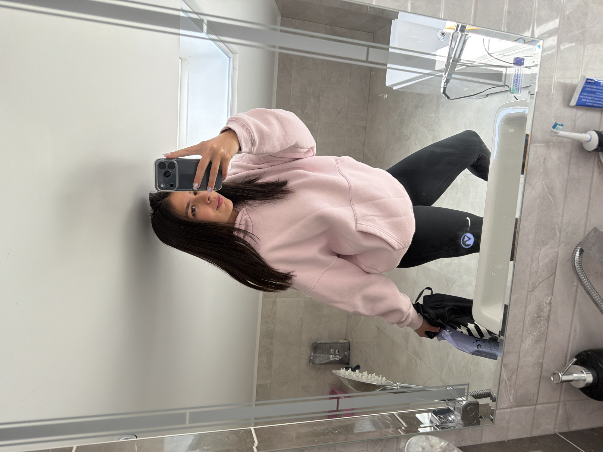 A young woman is reflected in a bathroom mirror, taking a selfie with a smartphone. She is wearing a light pink hoodie and black leggings. She is holding a black and white bag near the sink. The bathroom has light-colored walls and gray marble tiles. An electric toothbrush and a dispenser of toothpaste are mounted on the wall. The showerhead and faucet are chrome. The location is unknown.
