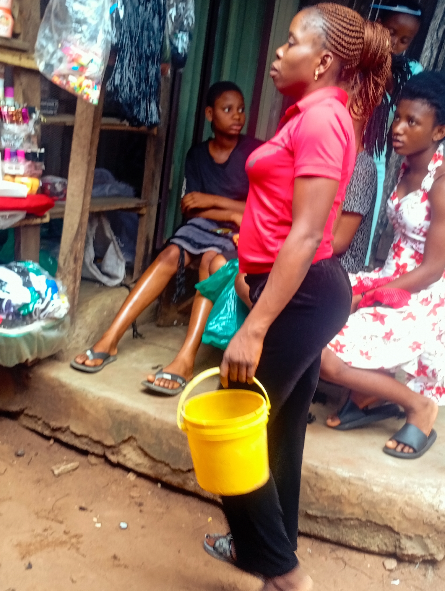 The image shows a group of young women in an outdoor setting, possibly a market or roadside stall. The woman in the foreground, on the right, is wearing a bright red polo shirt and black pants. She has her hair braided and pulled back in a ponytail and is holding a yellow bucket. Her posture suggests she is walking or standing.
To her left, another young woman is seated, wearing a black t-shirt and a striped skirt. She is looking to her right, with her legs crossed. Further to the right, another young woman is seated, wearing a white dress with a red floral pattern. She is looking towards the foreground. In the background, a fourth young woman is visible, wearing a black t-shirt and looking away from the camera.
The setting appears to be rustic, with a dirt ground and a raised concrete step or platform where some of the women are seated. There are shelves in the background displaying various items, some of which are wrapped in plastic. The overall lighting suggests it might be daytime. The expressions on the women's faces are difficult to discern clearly, but they appear to be engaged in their surroundings. There is no visible text in the image.
