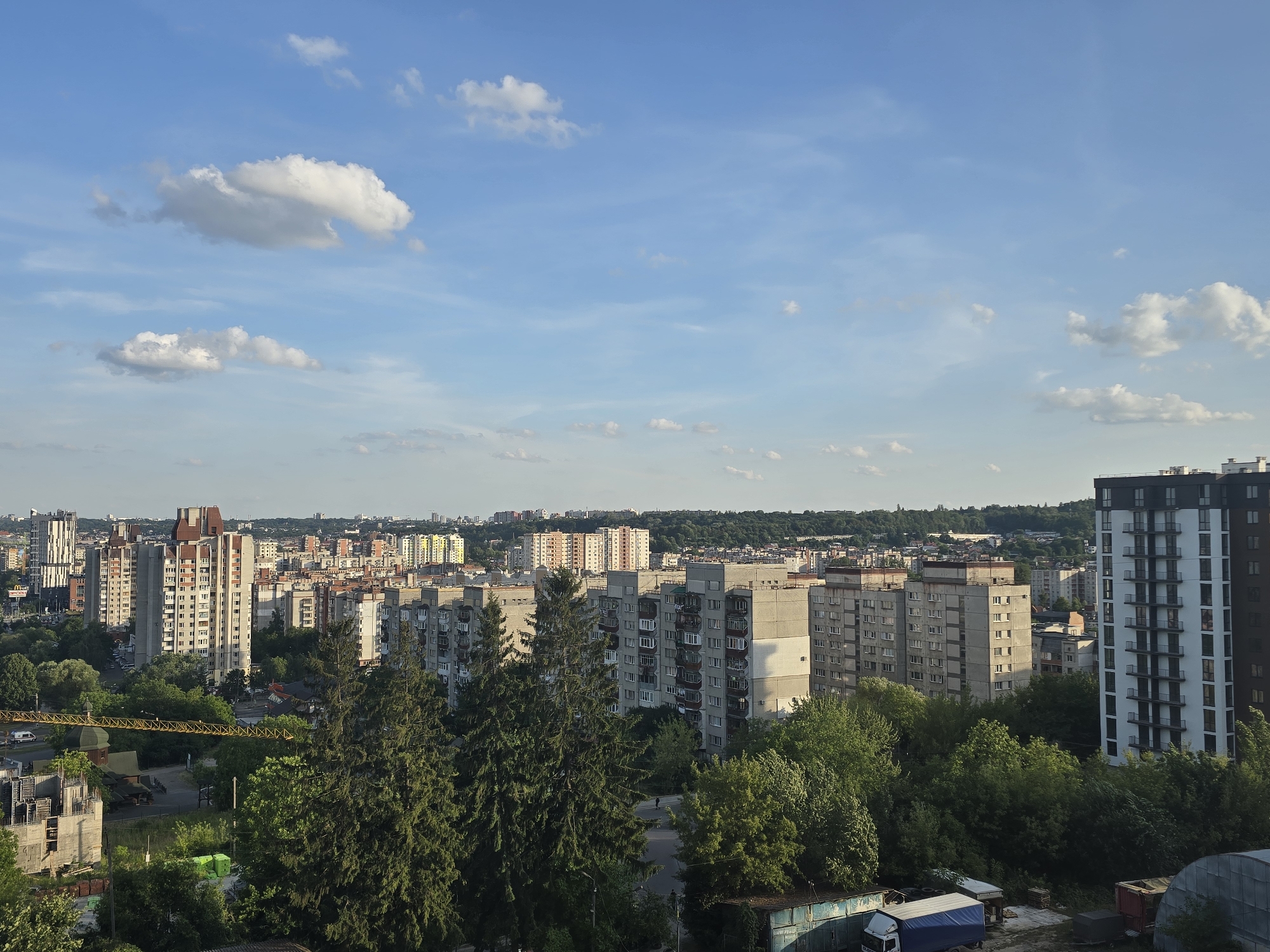 The image captures a panoramic view of a city district in Lviv, Ukraine, characterized by a mix of Soviet-era panel buildings and more modern high-rises. The sky is a clear blue, dotted with scattered white clouds, suggesting a pleasant daytime. In the foreground, lush green trees partially obscure a construction site with scaffolding and building materials. A yellow construction crane is also visible.
The mid-ground features a dense arrangement of apartment buildings, predominantly grey and beige, with visible balconies and windows. These buildings appear to be of similar architectural style, indicative of a planned residential area. Interspersed among the buildings are more trees and some visible roads with parked cars.
In the background, the cityscape extends further, with more residential buildings and some taller, more contemporary structures, including a distinctive white and black building on the right side of the frame. The horizon is lined with a gentle green slope of trees and hills. The overall impression is of a lived-in urban environment during daylight hours.
