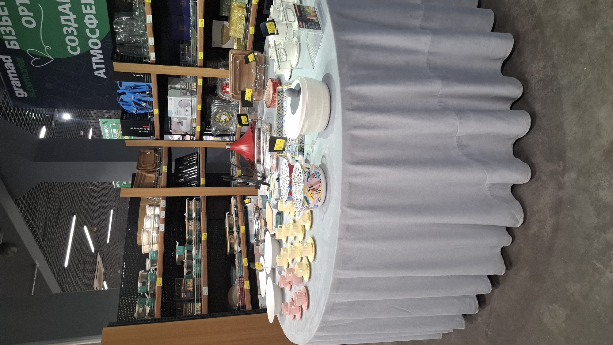 This media file captures an indoor scene, likely a supermarket or retail store, with a focus on kitchenware and home goods displayed on shelves and a central table. The overall lighting suggests an indoor environment, possibly during daylight hours given the bright illumination.
The primary subjects are various objects arranged for sale. On the shelves, we can see stacks of plates, bowls, and possibly cookware, with some items still in their packaging. The central table is adorned with an assortment of ceramic ware, including teacups and saucers in pastel colors like pink and yellow, as well as decorative bowls with colorful patterns. A large white pot or serving dish is also prominently featured on the table. Some of the items are marked with price tags or promotional labels.
The setting appears to be a modern retail space with a polished concrete floor. The background includes shelves stocked with more products, creating a sense of a well-organized store. There is no visible human presence in the frame, suggesting the photo was taken when the area was unpopulated or the individuals are out of frame.
Visible text includes signage on the left side of the image in Ukrainian. Text elements like "gramad" and "Supermarket" are discernible, indicating the brand or type of establishment. Another sign with green lettering reads "СКИДОК АКЦИИ" (discounts promotions). There are also smaller yellow price tags with numbers and symbols, typical of retail environments.
The arrangement of the goods on the table, particularly the neatly stacked teacups and saucers, suggests a deliberate display for customers. The overall impression is one of a commercial environment showcasing a variety of kitchen and dining accessories. The location context of Shymkent, Kazakhstan, is provided, but there are no specific visual cues within the image itself that definitively pinpoint this location, other than the Ukrainian text which might be present in some retail chains in the region.
