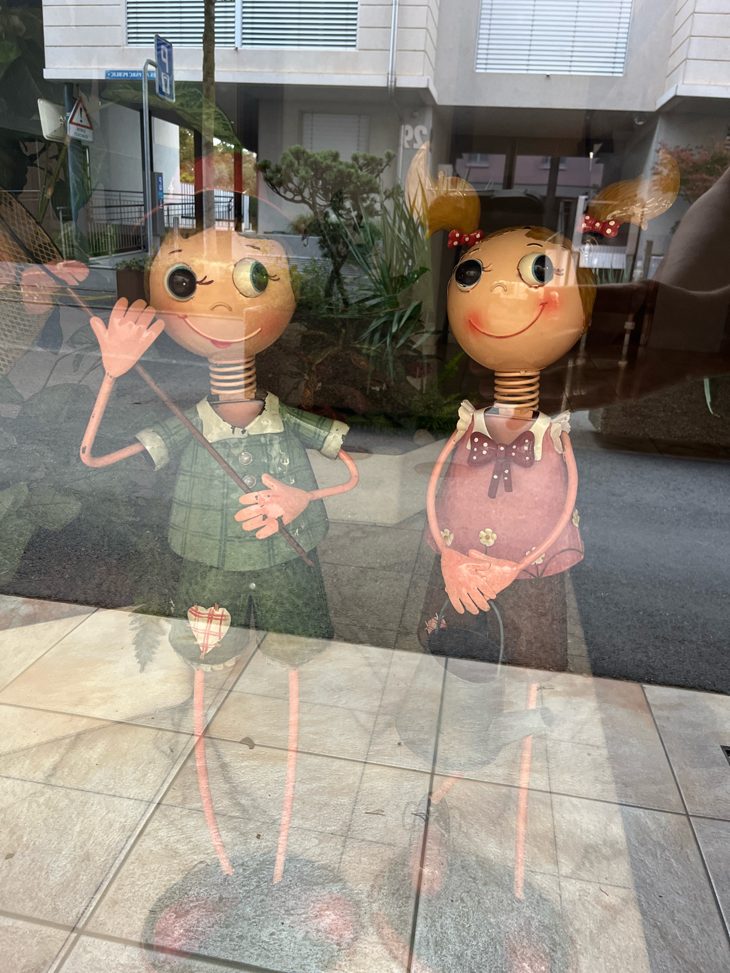 The image shows two whimsical metal figures, resembling children, displayed behind a reflective surface, likely a shop window, in Yverdon-les-Bains, Switzerland. The figure on the left has a round, tan head with large green and black eyes and a wide, red-lined smile. It wears a green, plaid shirt with three visible buttons and dark green pants adorned with a red and white checkered heart. Its left arm is raised, with its hand open as if waving, and it holds a thin brown stick. The figure on the right also has a round, tan head with similar large eyes and a smile, and two yellow pigtails tied with red polka-dot ribbons. It wears a pink dress with a white collar and a large maroon bow with white polka dots. Its hands are clasped together in front of its chest. The figures stand on circular bases, the reflections of which are visible on the tiled pavement outside. In the background, through the reflections, a building with horizontal white blinds and greenery can be seen. A blue traffic sign with a white 'P' symbol and a yellow triangular warning sign are also visible on a pole. The overall scene is brightly lit, suggesting daytime.