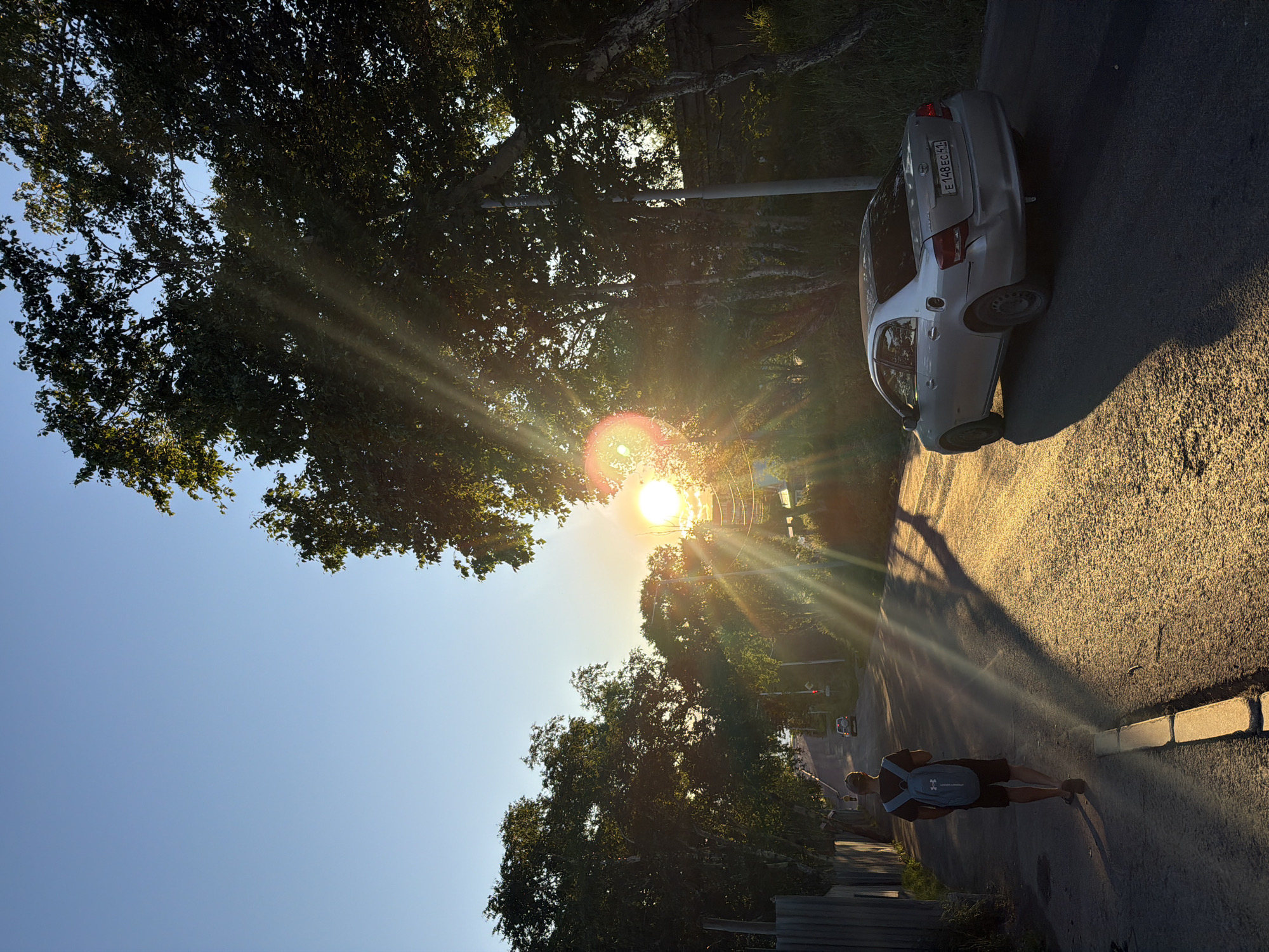 In Petropavlovsk-Kamchatsky, Russia, a young person walks down a paved road lined with trees. The sun is setting, casting long shadows and creating a golden glow on the asphalt. A silver sedan is parked on the right side of the road. The sky is clear blue, with the sun partially obscured by trees. The scene appears to be a quiet residential street.