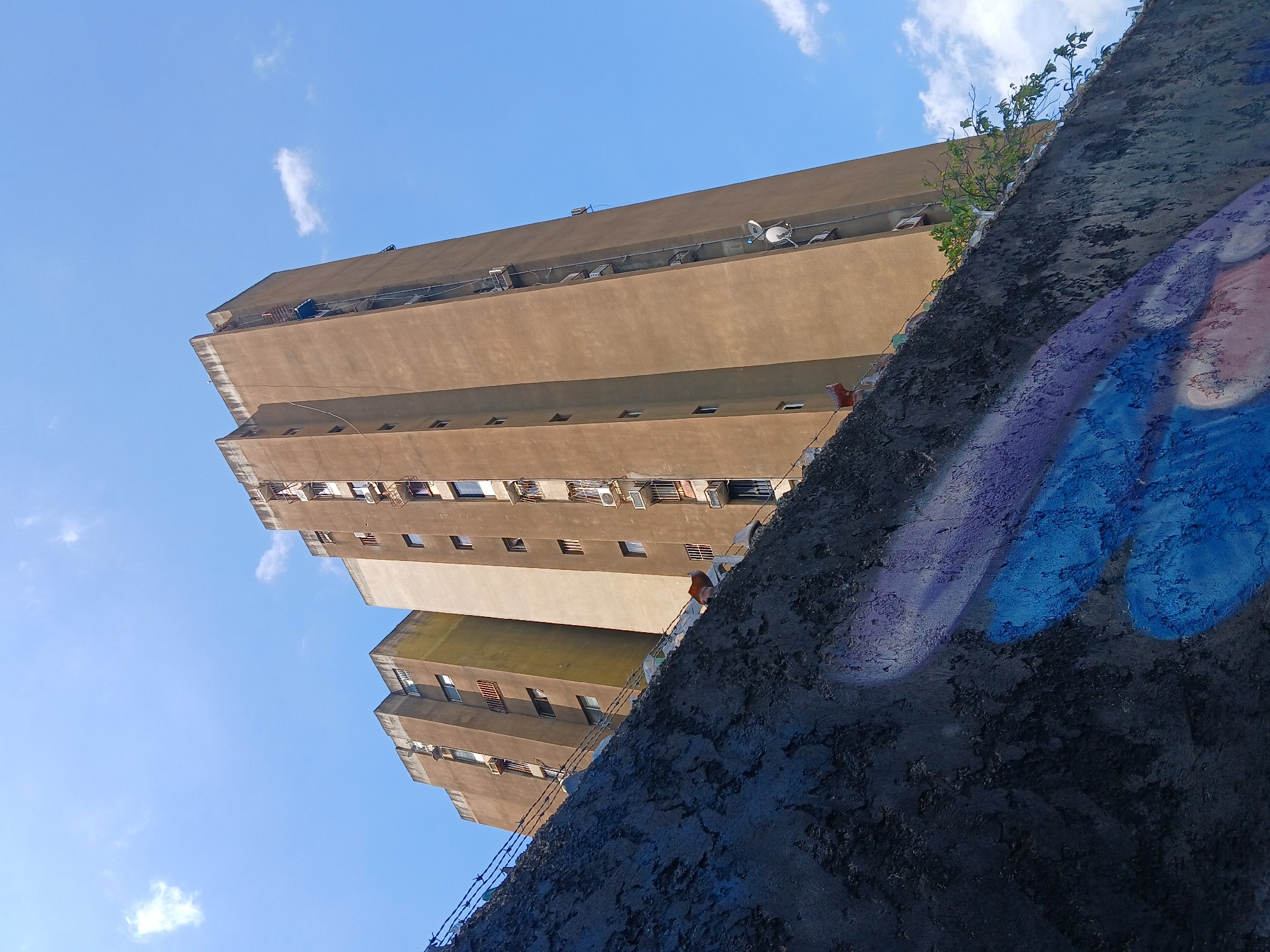The image displays an upward-looking perspective of a tall, multi-story building, likely an apartment block, under a clear, bright blue sky with a few wispy white clouds. The building has a brownish-tan facade, characterized by numerous small rectangular windows and some sections with small balconies or utility spaces. Several white satellite dishes are mounted on the upper levels or rooftops.
In the foreground, a dark, textured concrete wall diagonally occupies the right side of the frame. This wall is decorated with abstract graffiti in shades of blue and purple. Green foliage grows from the top edge of this wall, and barbed wire is visible along its upper boundary in some areas. The scene depicts a static urban landscape in Ciudad Guayana, Venezuela, illuminated by bright daylight, suggesting sunny weather conditions. No people or specific activities are present. There is no discernible text within the image.