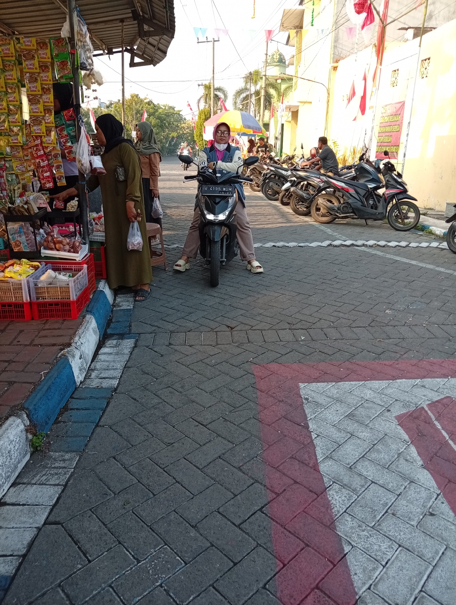 In Bligo, Indonesia, a street-side market bustles with activity under a canopy. A vendor, wearing a dark hijab and an olive green dress, gestures towards shelves stocked with packaged snacks. Beside her, another woman in a light brown hijab stands with a plastic bag. In the foreground, red and white plastic crates hold various goods.
The street is paved with interlocking gray bricks, bordered by a curb painted in alternating blue and white. Several motorcycles are parked along the street, with a woman in a light pink hijab and glasses seated on one in the center, facing the camera. The background reveals buildings, trees, and flags, suggesting a festive or commemorative occasion. The lighting indicates it is daytime, with sunlight casting shadows.