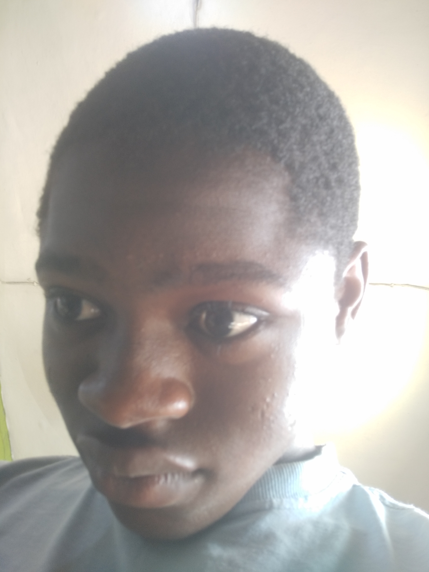 A close-up photo of a person with dark skin and short, dark hair. The person is wearing a blue-gray shirt. The background is a light-colored wall. The location in Shangisha, Nigeria, is not discernible from the image.