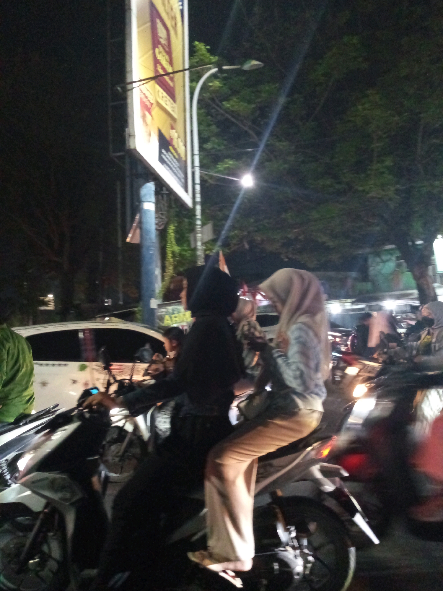 The image captures a night scene in the city of Bekasi, Indonesia, likely on a street with trees and a billboard. Two individuals are prominently featured on a scooter in the foreground. The person in the front, wearing a black hijab and dark clothing, is operating the scooter. The person in the back, also wearing a hijab and a patterned top with light-colored pants, is seated behind them. Their bodies are angled towards each other, suggesting interaction. A white car is visible behind the scooter. Other scooters and indistinct figures are present in the background, illuminated by streetlights and the scooter's headlights. The overall atmosphere is that of a busy urban environment at night.