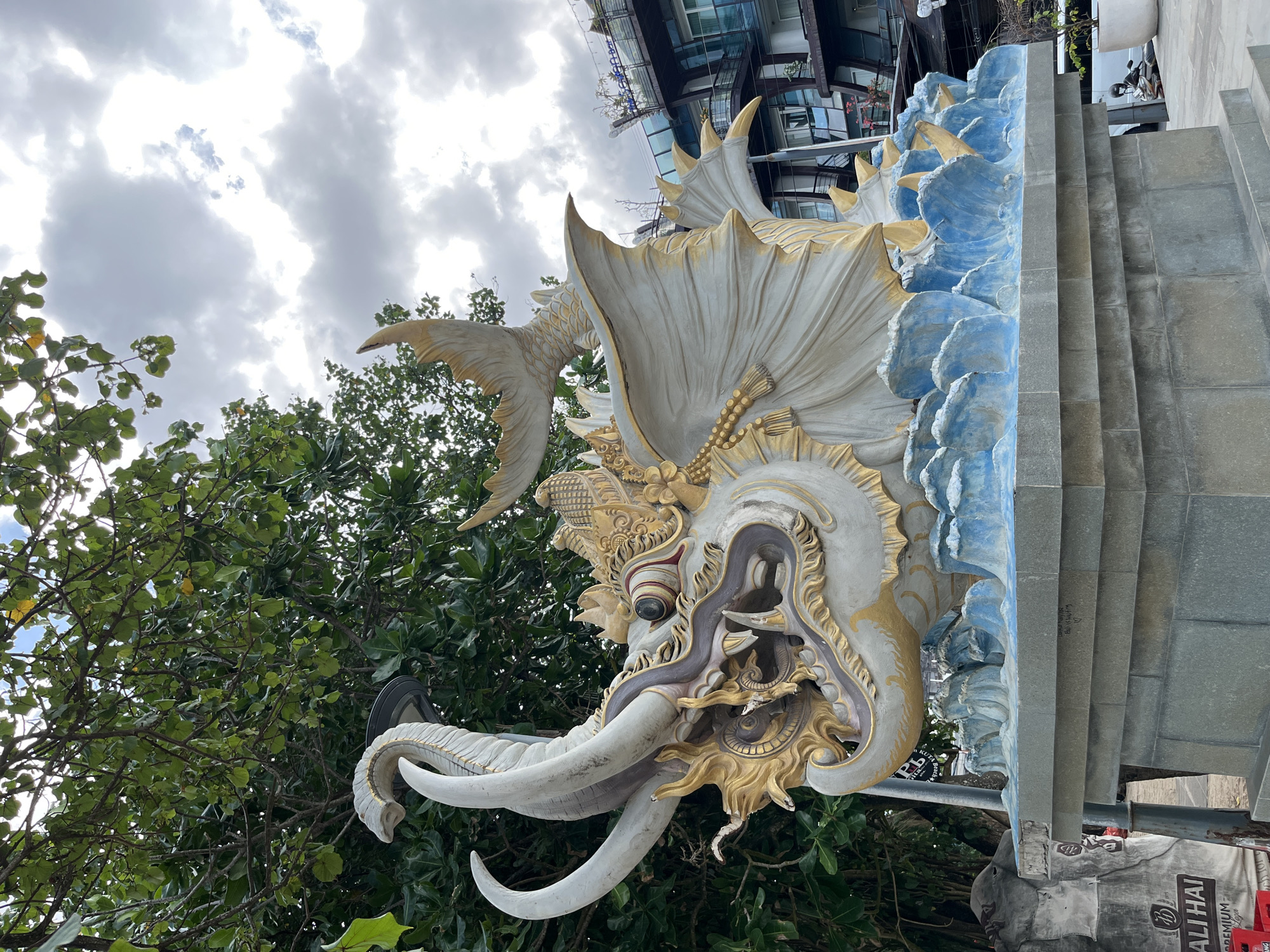 This image shows a close-up of a large, ornate statue of a mythical creature, possibly a dragon or elephant-like being, with prominent tusks and intricate golden detailing. The statue is predominantly white and gold, with accents of blue designed to resemble waves or water. It appears to be part of an architectural feature, possibly a gate or entrance, with stone steps visible to the right.
The statue is set against a backdrop of lush green trees and a cloudy sky. In the upper right portion of the image, a modern building with glass windows is visible, suggesting an urban or developed setting. The presence of the statue and the building hints at a location that blends traditional or religious art with modern architecture.
The lighting suggests it might be daytime, though the overcast sky could indicate a cloudy day or late afternoon. There are no people visible in the frame.
Visible text includes "ULI HAI" on a banner at the bottom right of the image, and possibly other unreadable text on a poster below the steps. The location context provided is Kuta, Indonesia.