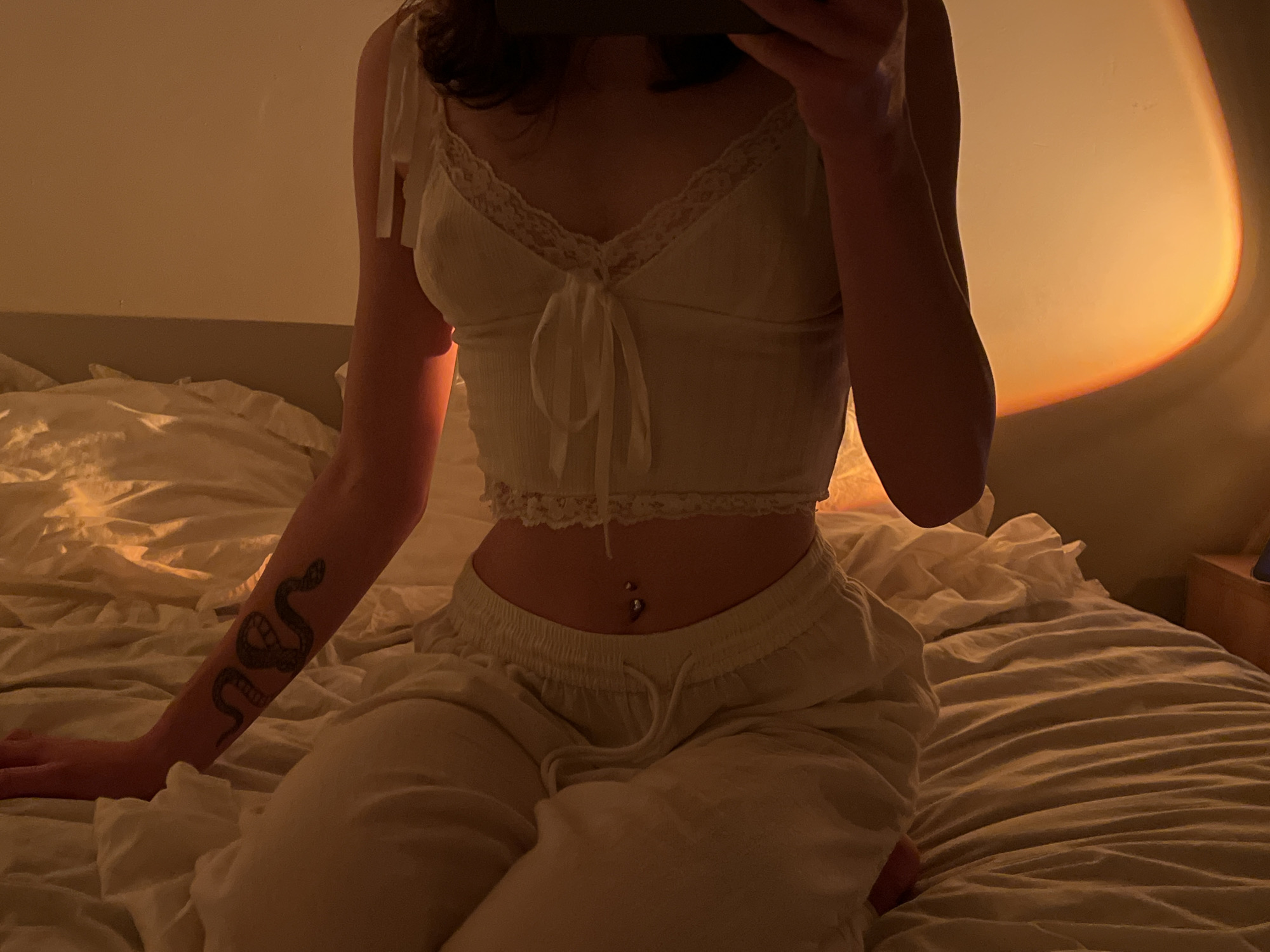 The image shows a person sitting on a bed in a dimly lit room. The person is wearing a white camisole and light-colored pants, and has a snake tattoo on their left arm. A belly button piercing is visible. The room appears to be a bedroom, with rumpled white bedding. A warm, orange light is cast on the wall behind the person, possibly from a sunset lamp or artificial lighting. The location is not identifiable as Ternopil, Ukraine, or any specific city.