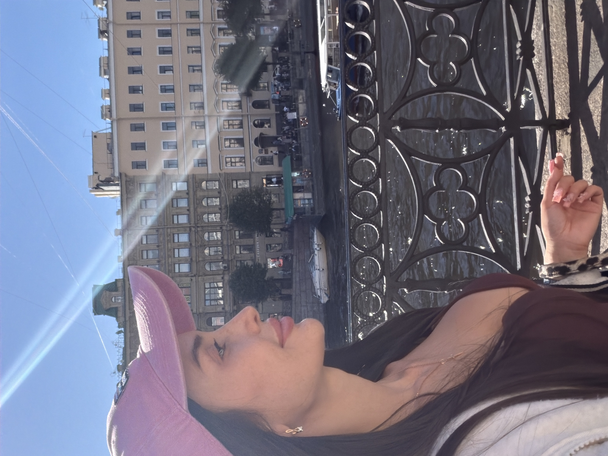 A profile portrait of a woman wearing a pink baseball cap and a dark maroon top is captured against the backdrop of Saint Petersburg, Russia. The woman is looking to her left, with her long, dark hair cascading down her shoulders. She is adorned with a delicate necklace and a small, sparkly earring.
The setting appears to be a waterfront promenade. In the background, several multi-story buildings with classical architecture line the canal. A dark metal railing, decorated with circular and clover-shaped motifs, runs along the foreground, separating the viewer from the water. Several boats are visible on the canal, including a white motorboat and a larger tour boat. The sky is a clear blue, with some contrails from aircraft visible. Sunlight streams into the image, creating lens flare and casting shadows on the ground. The overall atmosphere is bright and sunny, suggesting a pleasant day in the city.