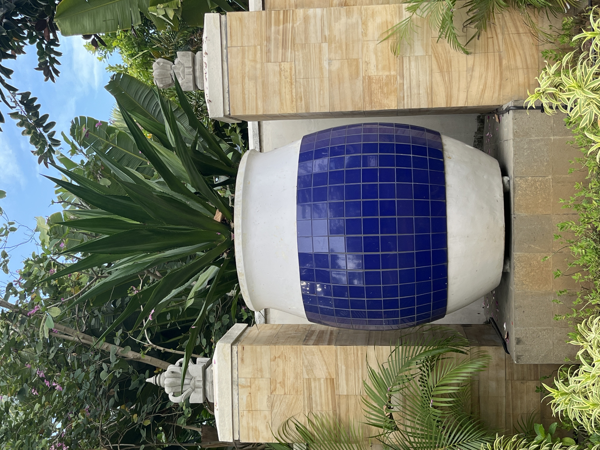 In Kuta Selatan, Indonesia, a large, white ceramic planter with a blue tiled midsection stands prominently, housing a vibrant green agave plant. The planter rests on a tiered stone base, flanked by beige tiled pillars topped with decorative stone finials. Lush foliage, including palm fronds and trees with small pink flowers, surrounds the planter, creating a tropical ambiance. The scene is set against a backdrop of a bright blue sky.