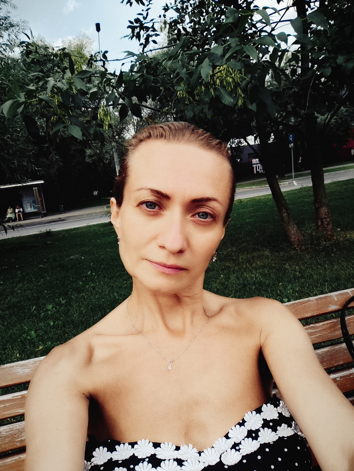 A woman with brown hair pulled back is sitting on a park bench, facing the camera. She is wearing a black strapless dress with white polka dots and a white floral trim. A delicate silver necklace with a small pendant is visible on her neck. Her eyes are blue and she has a neutral expression. Behind her, there is a grassy area, trees, a street, and a bus stop with two people sitting on a bench. The setting appears to be a city park, likely in Moscow, Russia, given the architecture and typical urban park elements visible. The lighting suggests it is daytime.