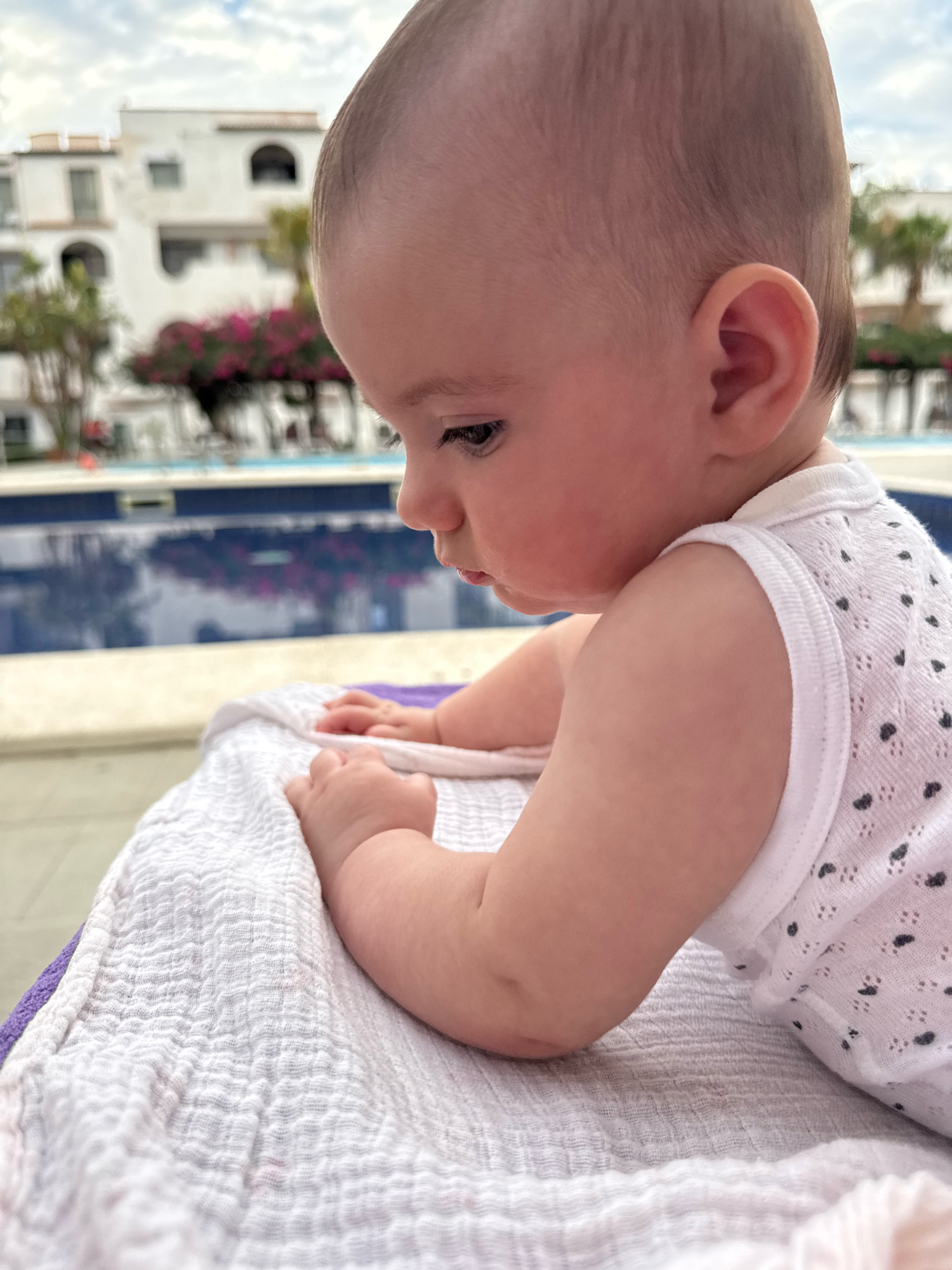 A baby is lying on a textured white blanket and a purple towel, with their head turned to the right and looking down. The baby is wearing a white sleeveless top with a pattern of small black dots. In the background, there is a swimming pool with blue water and reflections of buildings and trees. The buildings appear to be white, multi-story structures with arches and balconies, consistent with the architecture found in Isola delle Femmine, Italy, a coastal town known for its residential buildings and vacation accommodations. The sky above is partly cloudy, with a mix of blue sky and white clouds. The overall scene suggests a relaxed outdoor setting, possibly a hotel or residential complex with a pool area.