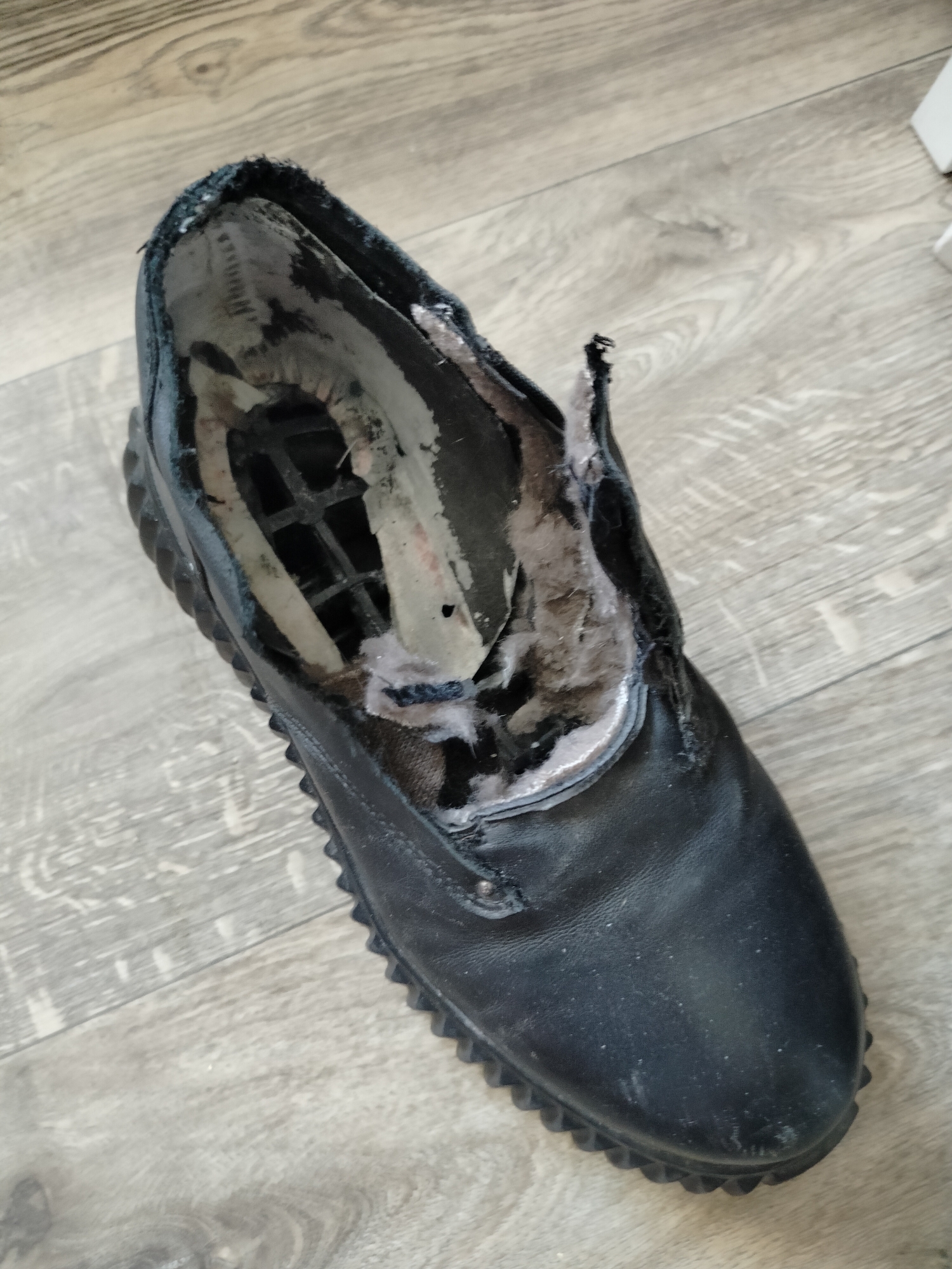 A single, black, worn-out shoe lies on a light gray wood-grain floor. The shoe's opening is torn and frayed, revealing the inner lining and some of the shoe's structure. The sole of the shoe is thick and black with a serrated edge. The floor appears to be indoors, possibly in a residential or commercial setting. There are no people or any other objects in the immediate vicinity of the shoe. The lighting suggests an indoor environment with overhead or side lighting. The scene is devoid of any context that would place it in Novosibirsk, Russia.