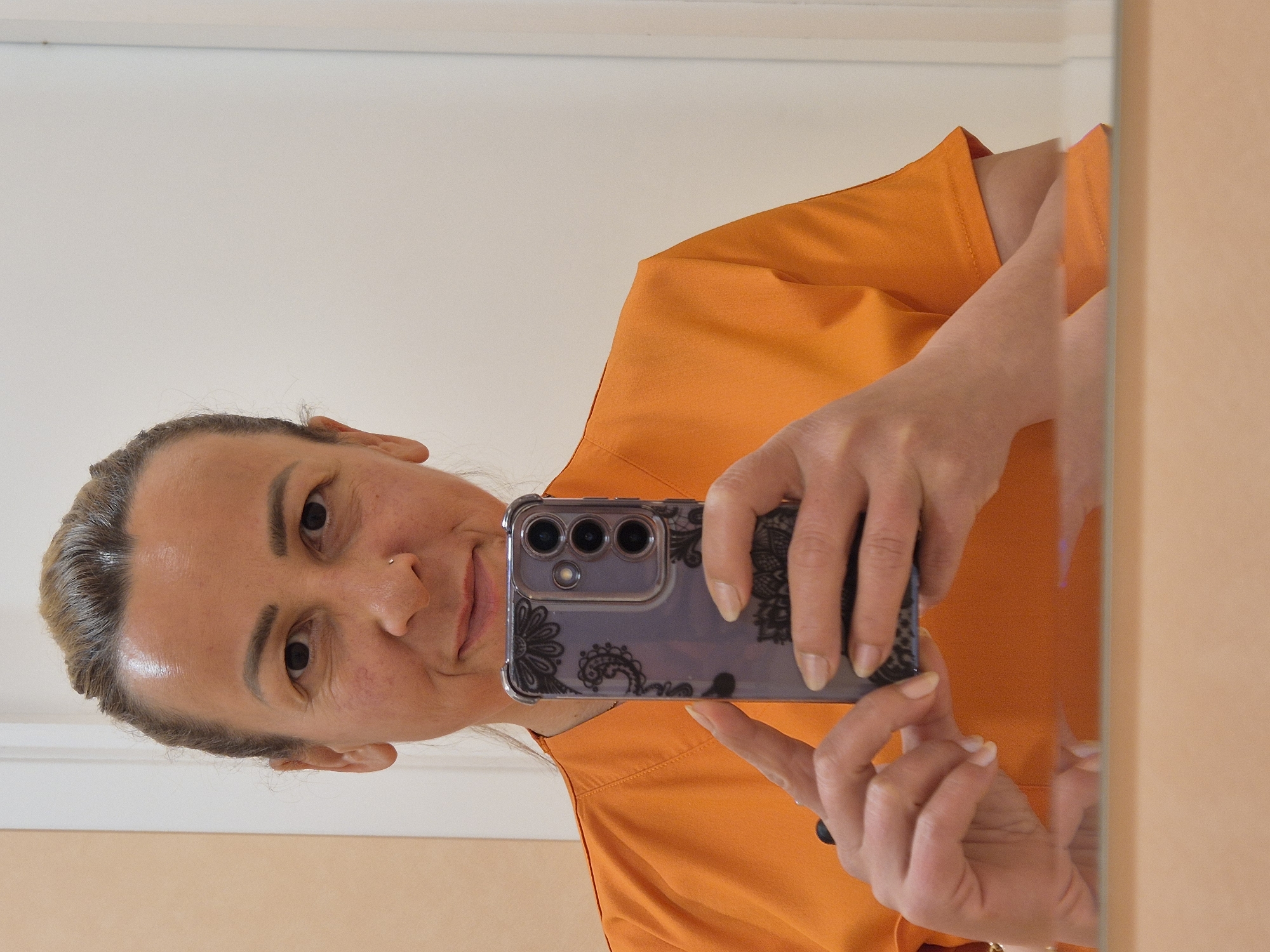 A woman, appearing to be in her 30s or 40s, with blonde hair pulled back, takes a mirror selfie. She is wearing an orange scrub top and has a small nose ring. Her face is turned towards the camera, and she is holding a smartphone in her right hand, with her left hand resting on the phone. The phone case has a black lace pattern. The background shows a wall and a doorframe, suggesting an indoor setting, possibly a changing room or bathroom. The reflection in the mirror shows her from the chest up, and also a blurry reflection of her lower body.