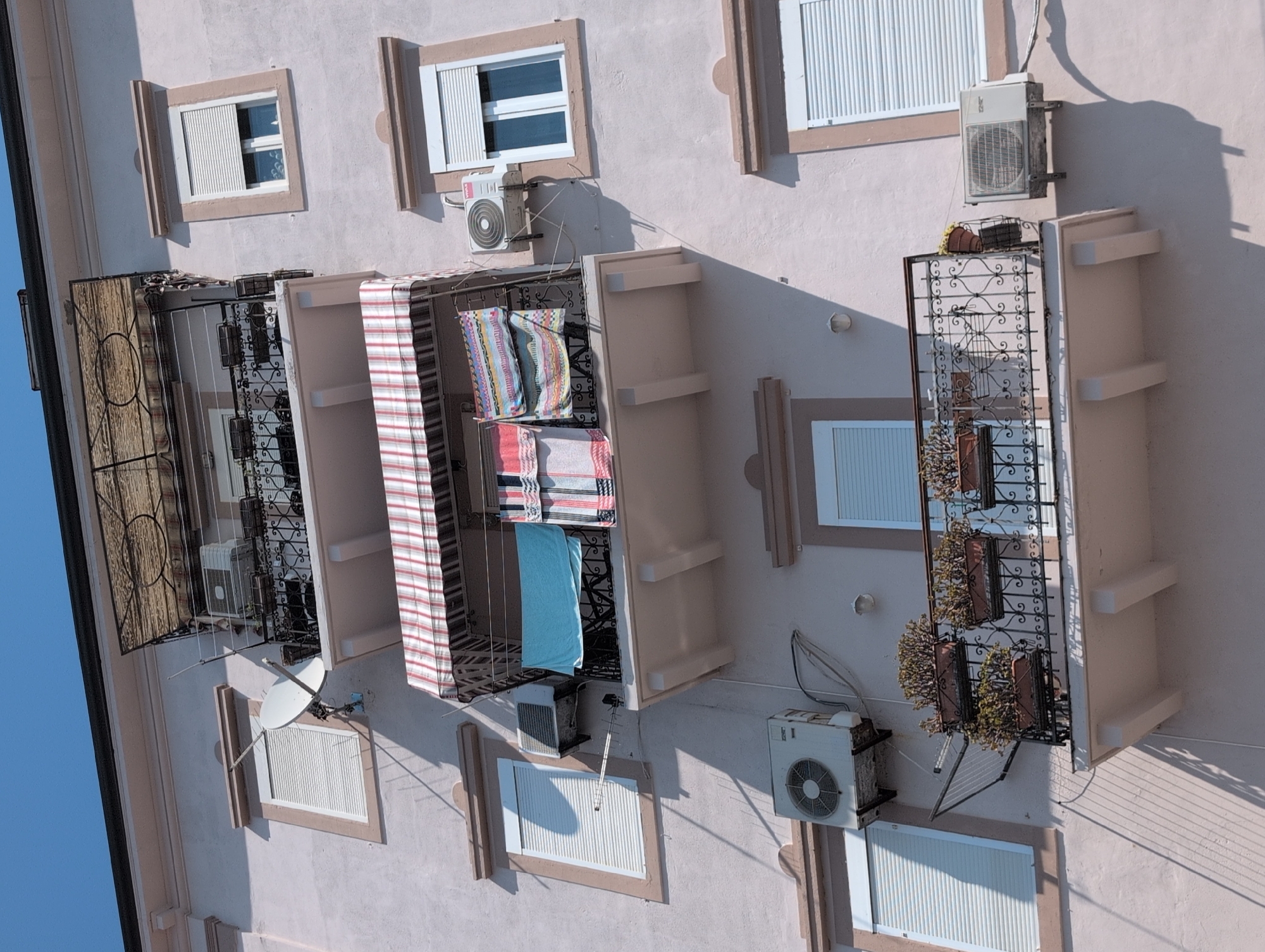 This is a medium shot, eye-level, exterior, daytime photograph of a light pink building in the city of Grad Rijeka, Croatia. The building features multiple balconies with various items on them, including clothes drying on a line, planters with vegetation, and air conditioning units. One balcony has a striped awning. A satellite dish is visible on the left side of the building. The sky is clear and blue.