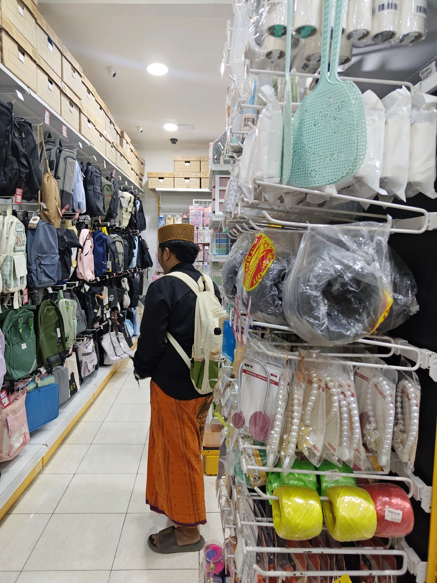 The image captures a person in a black shirt, an off-white backpack, and an orange sarong, wearing a traditional cap, browsing through a retail store that appears to specialize in bags and various household items. The store's interior is brightly lit, with rows of shelves stacked with merchandise. On the left, numerous backpacks in different colors and styles are displayed on shelves. To the right, a rack holds an assortment of packaged goods, including what look like fly swatters, rolls of string in yellow and red, and strings of pearl-like beads. The flooring is tiled with a light-colored pattern. The overall environment suggests a typical retail setting, likely within a commercial area in Situbondo, Indonesia, given the sarong and cap which are common attire in the region. The person's gaze is directed towards the bags on the left, indicating they are in the process of shopping.